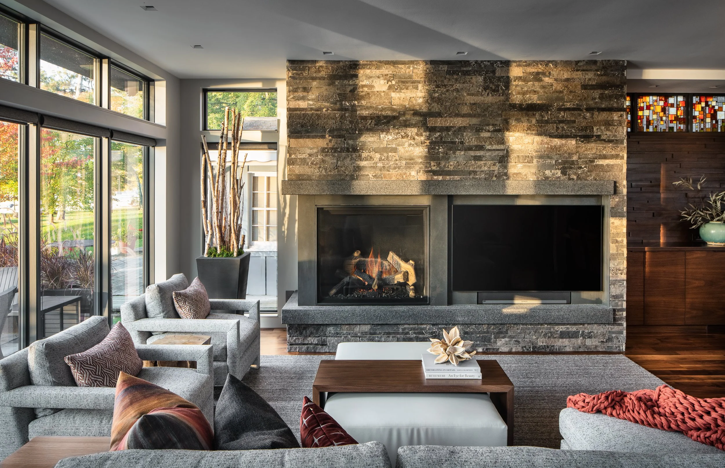 Modern living room with a large stone fireplace, gray furniture with decorative pillows, and floor-to-ceiling windows showing a view of trees outside. There is a wooden table and some decorative books and plants.