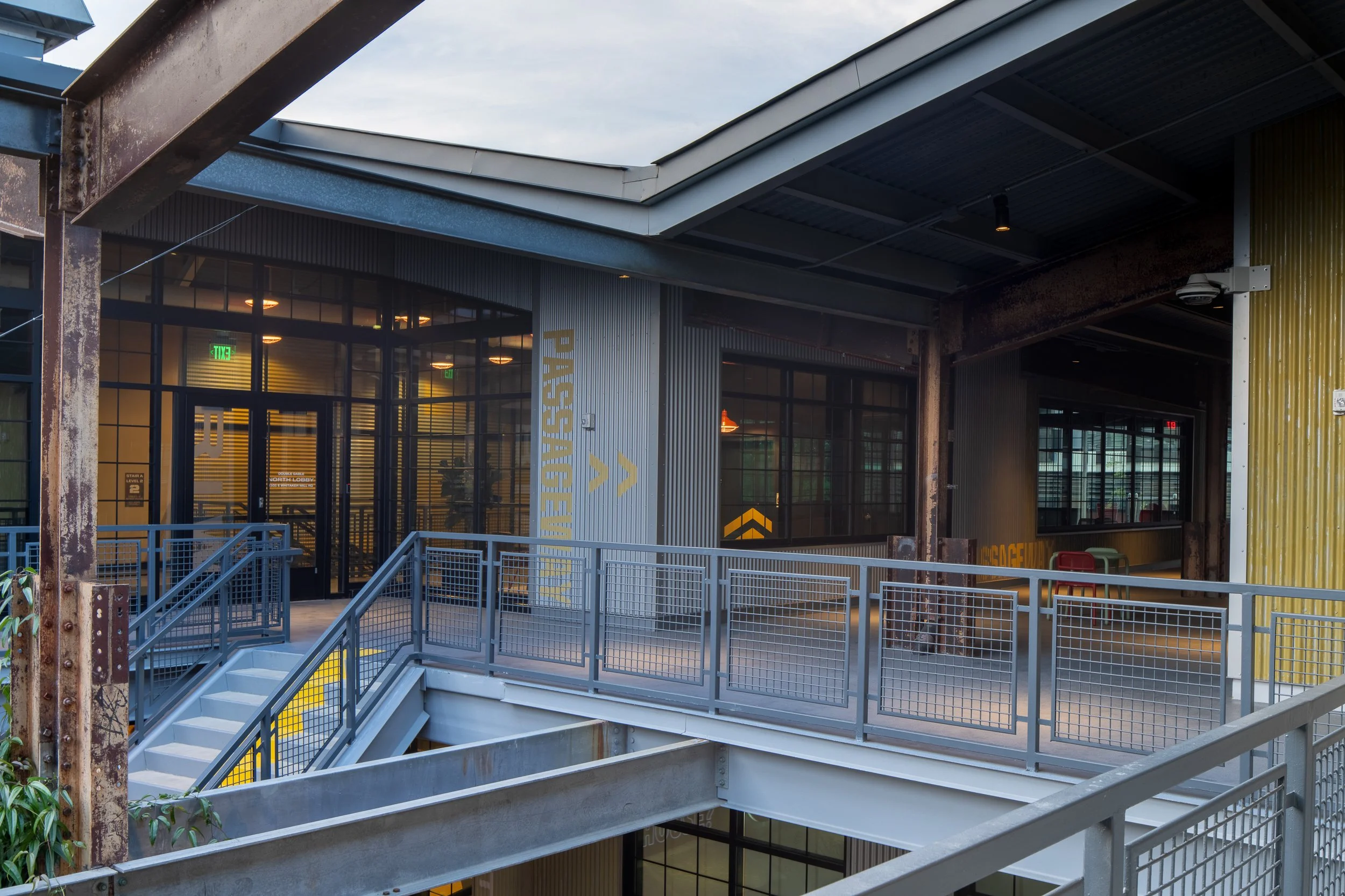 Second-floor walkway with mesh railing, original rusted steel column on the left, yellow painted PASSAGEWAY graphic on the corrugated wall, and warm interior light spilling through industrial windows at dusk.