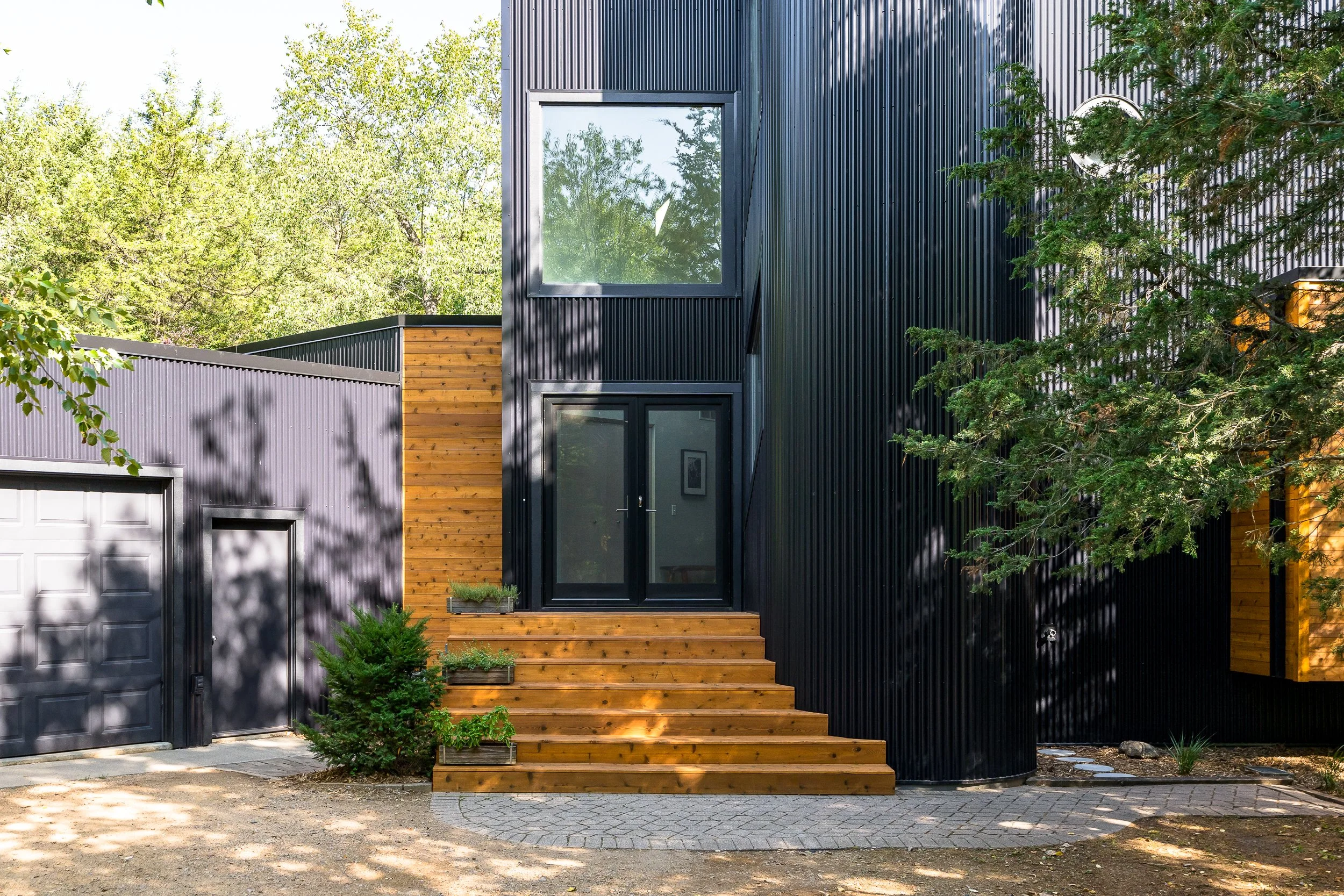 Black modern home exterior with vertical siding, wood entry steps, and shaded trees.