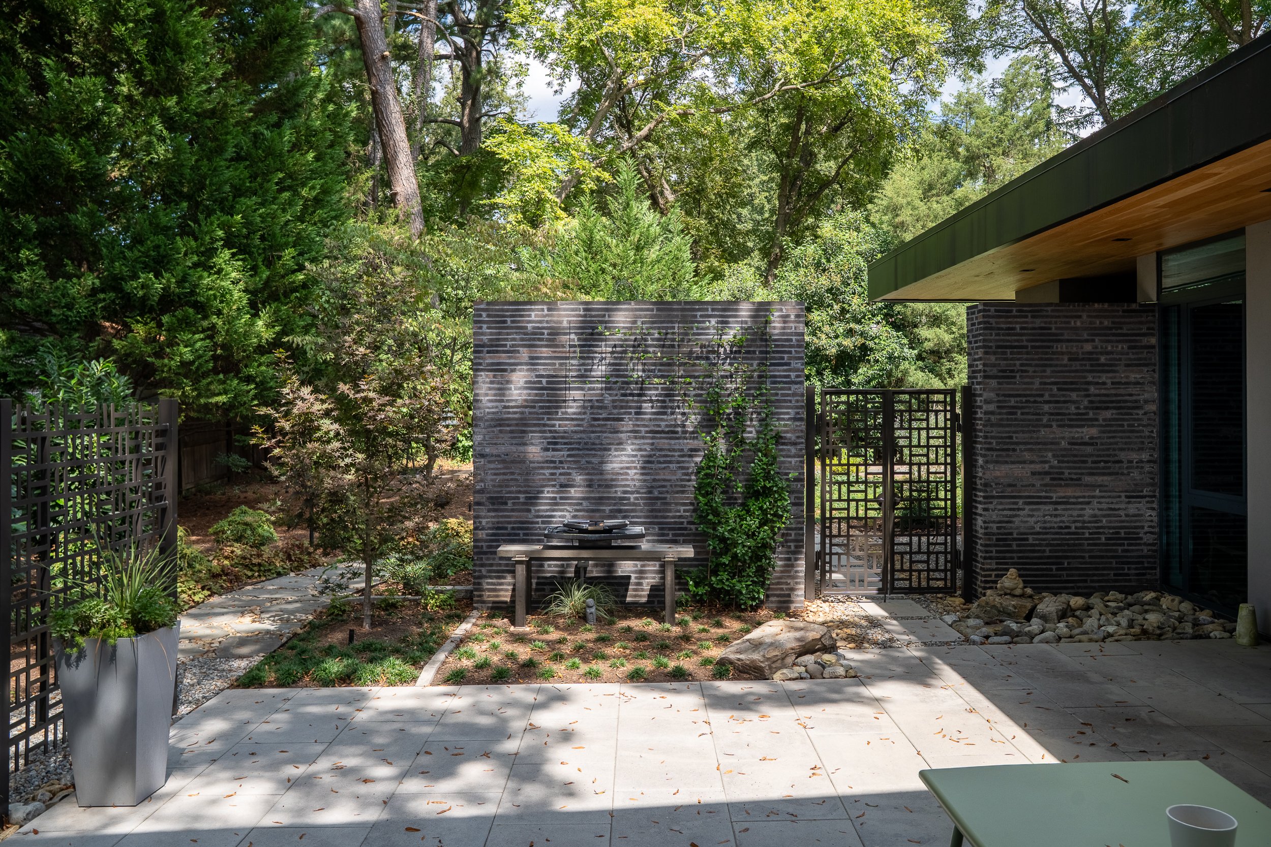 A paved courtyard bordered by dark brick walls contains a bench, potted planting, and a gate opening toward dense greenery.