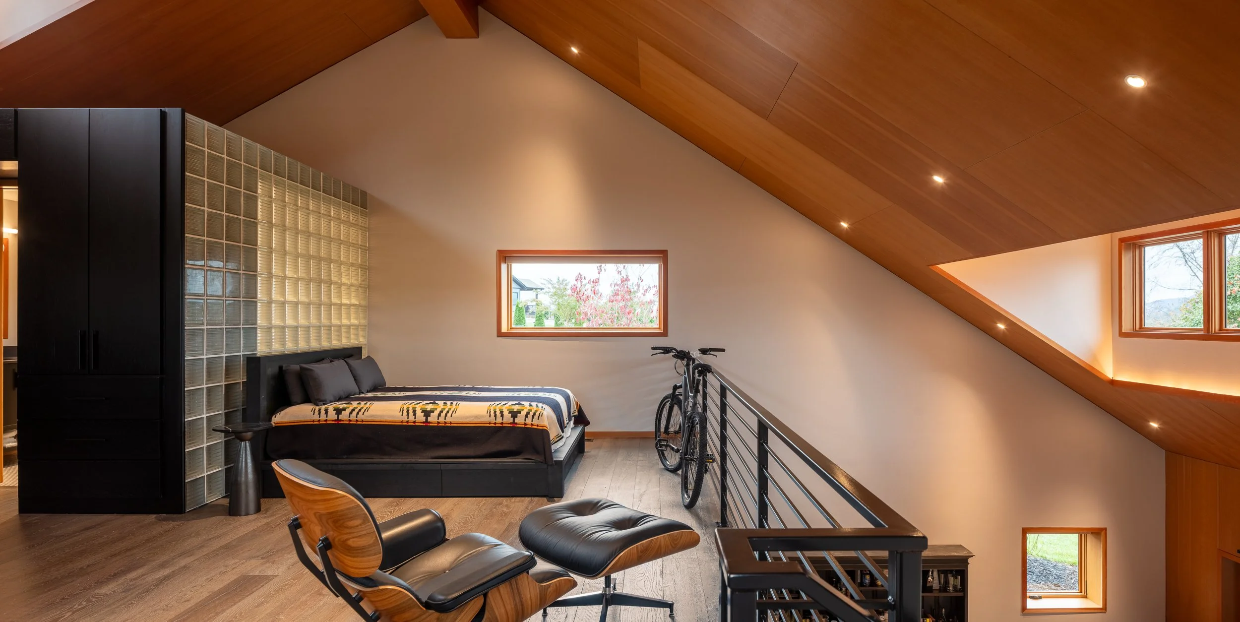 Loft bedroom with bicycle, black railing, contemporary lounge chair, and angled wood ceiling.