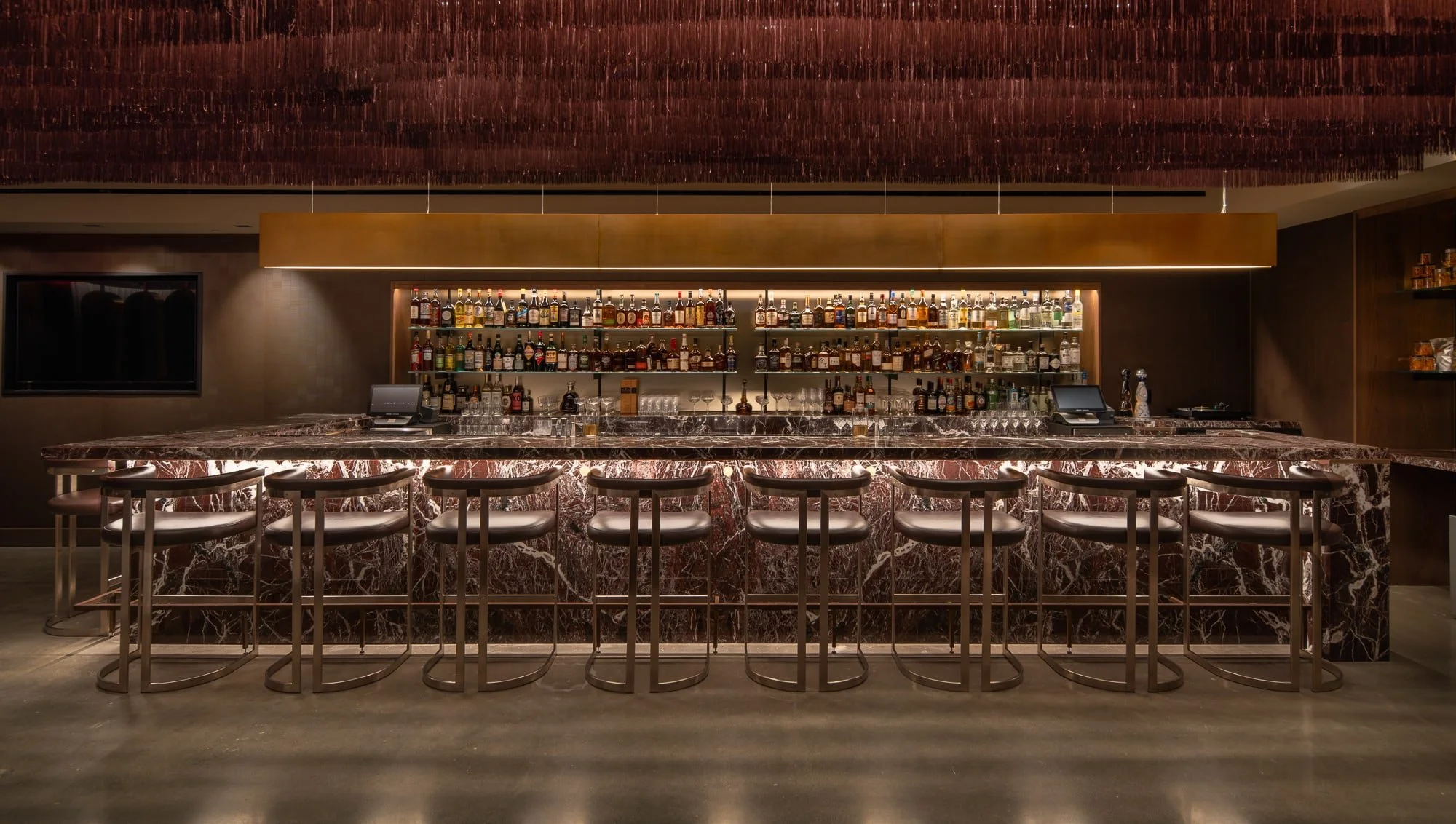 Backlit marble bar with leather stools, illuminated bottle shelves, bronze accents, and a suspended burgundy ceiling feature.