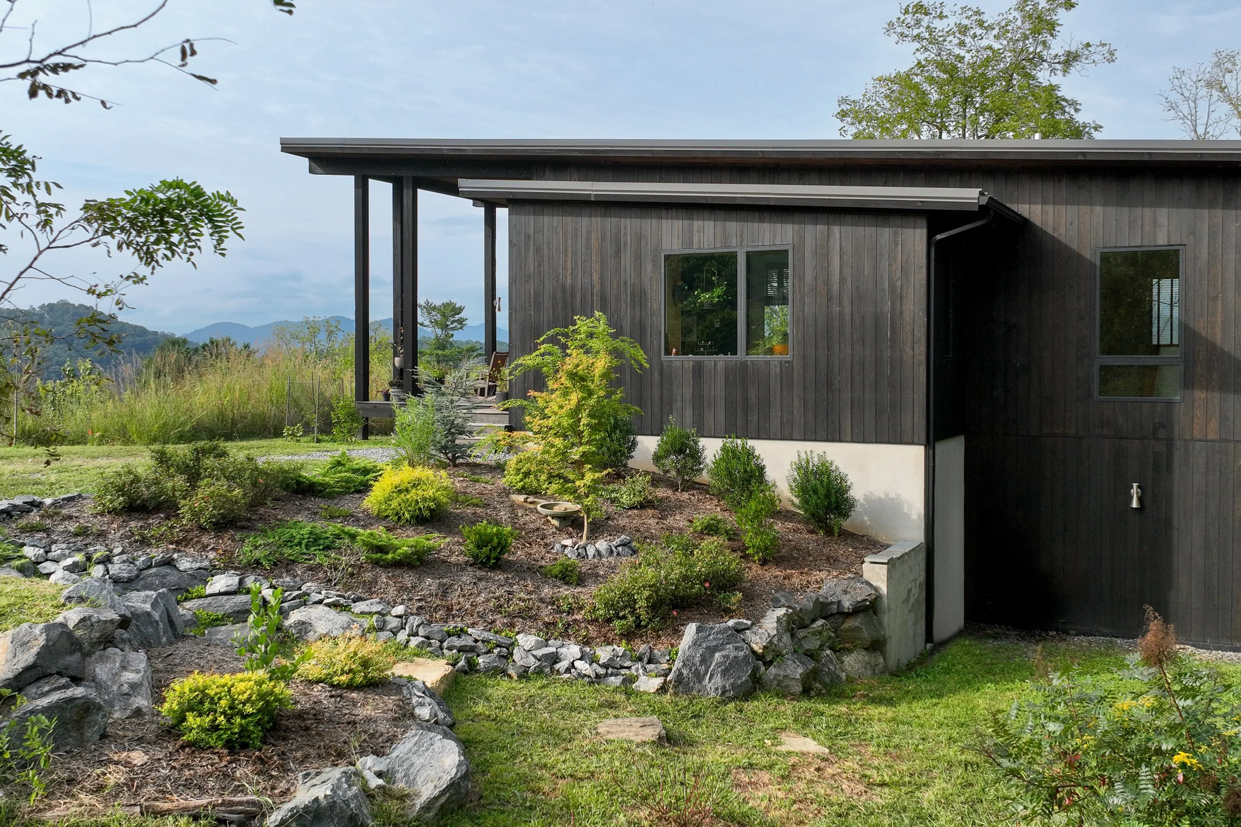 A modern house with dark wooden exterior walls and several large windows, surrounded by lush landscaping and greenery, with a mountainous landscape in the background.