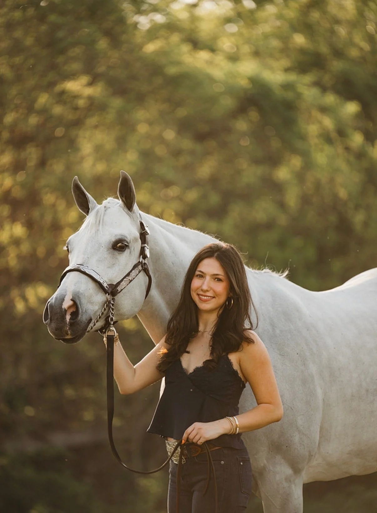 Person walking a brown horse along a gravel path in a green, sunny landscape with trees and a white fence.
