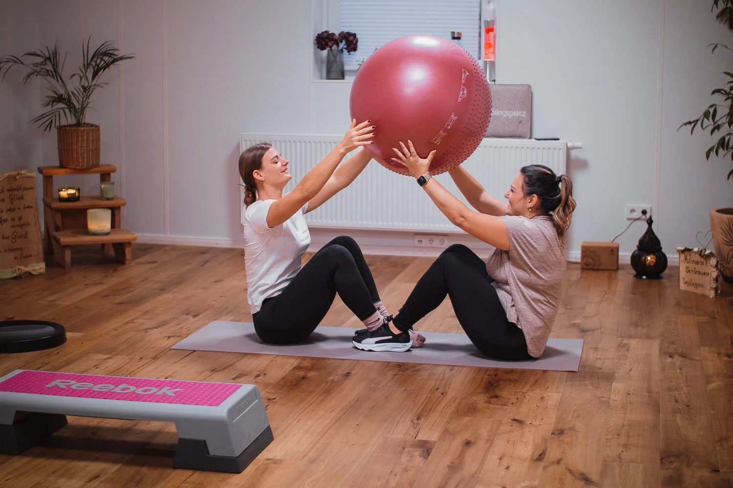 Zwei Frauen auf Gymnastikmatten, die einen großen, roten Gymnastikball halten. Der Raum hat Holzboden, Pflanzen und eine Bank mit Reebok-Logo. Es gibt dekorative Kerzen und Regale im Hintergrund.