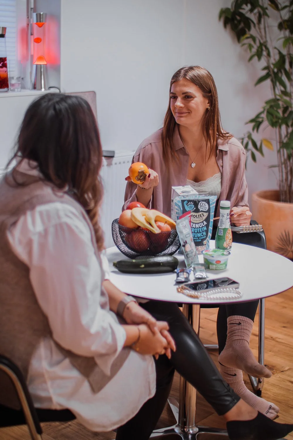 Zwei Frauen sitzen an einem Tisch und unterhalten sich. Auf dem Tisch befinden sich Obst, Verpackungen von Lebensmitteln und ein Smartphone. Eine Frau hält eine Frucht in der Hand. Im Hintergrund sind eine Lavalampe und eine Pflanze zu sehen.