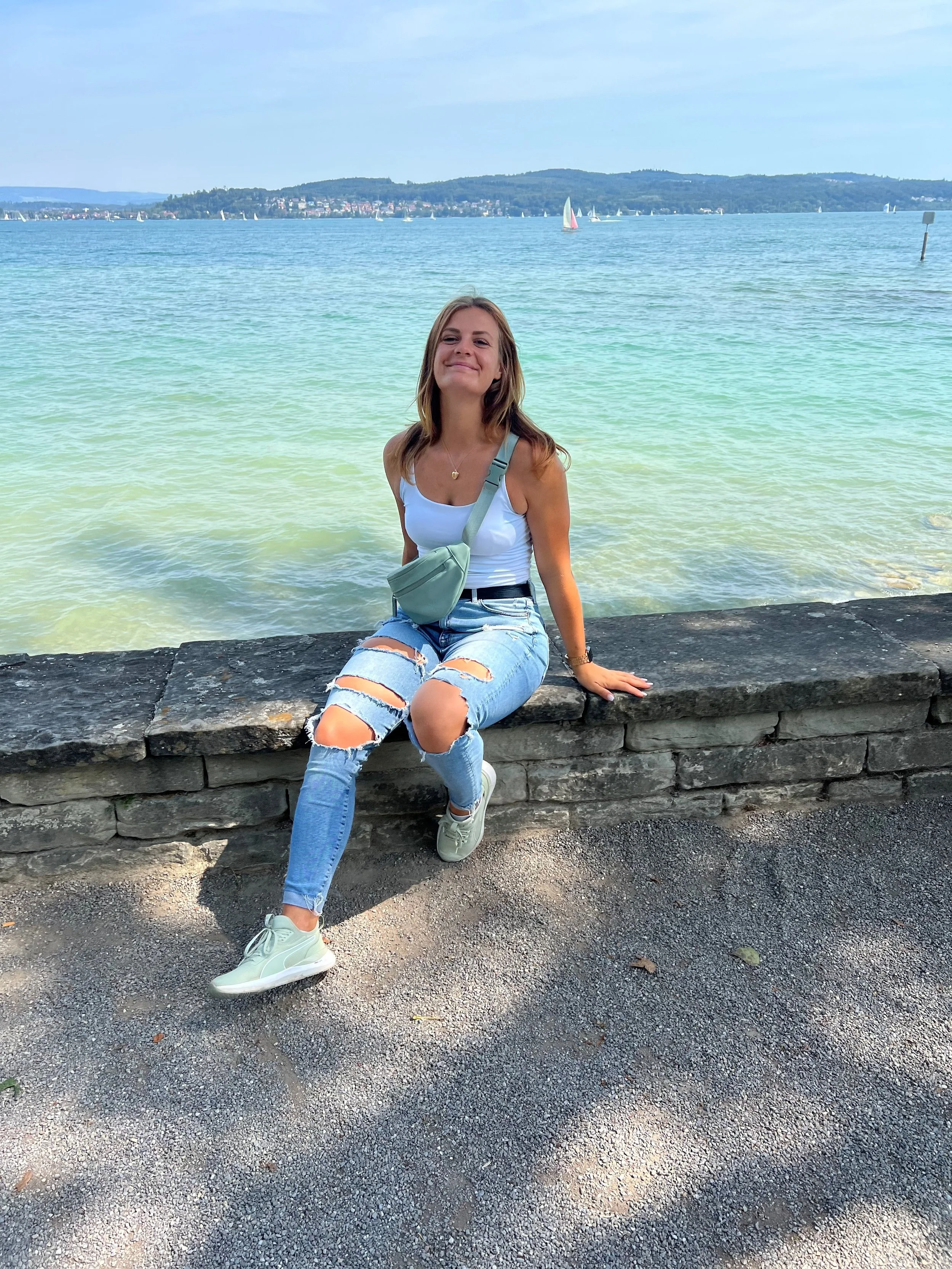 Frau sitzt auf einer Steinmauer am See mit Segelbooten im Hintergrund.