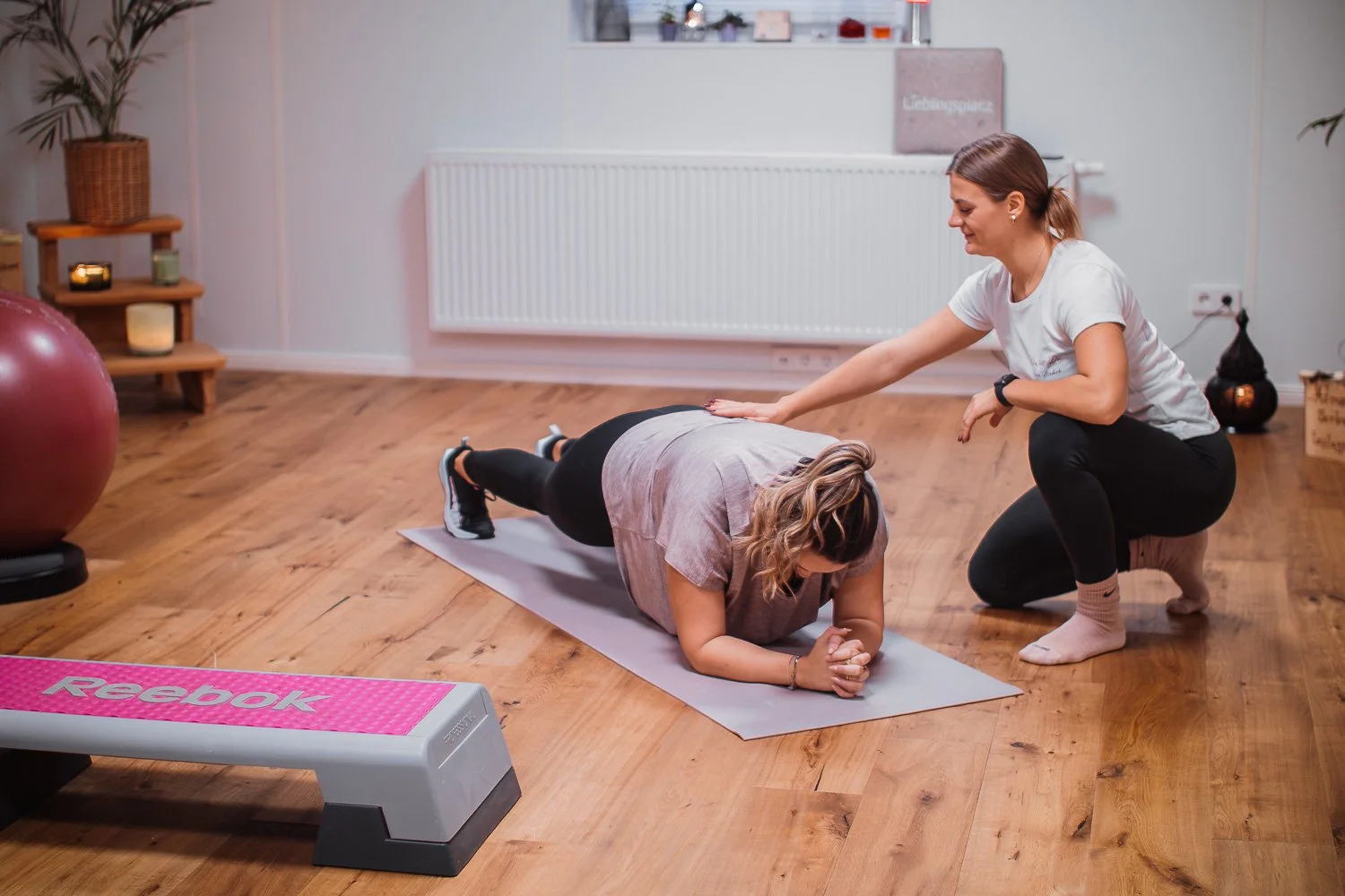 Zwei Frauen in einem Fitnessstudio, eine macht eine Plank-Übung auf einer Yogamatte und die andere gibt Anweisungen; im Raum sind ein Gymnastikball und ein Stepper.