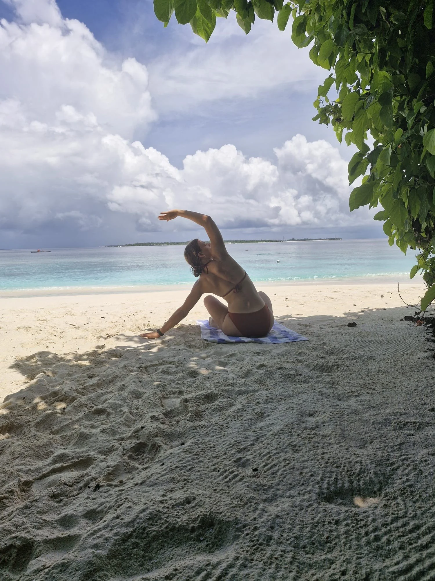 Person am Strand, macht Yoga, Blick auf das Meer, blauer Himmel, Wolken, Bäume.