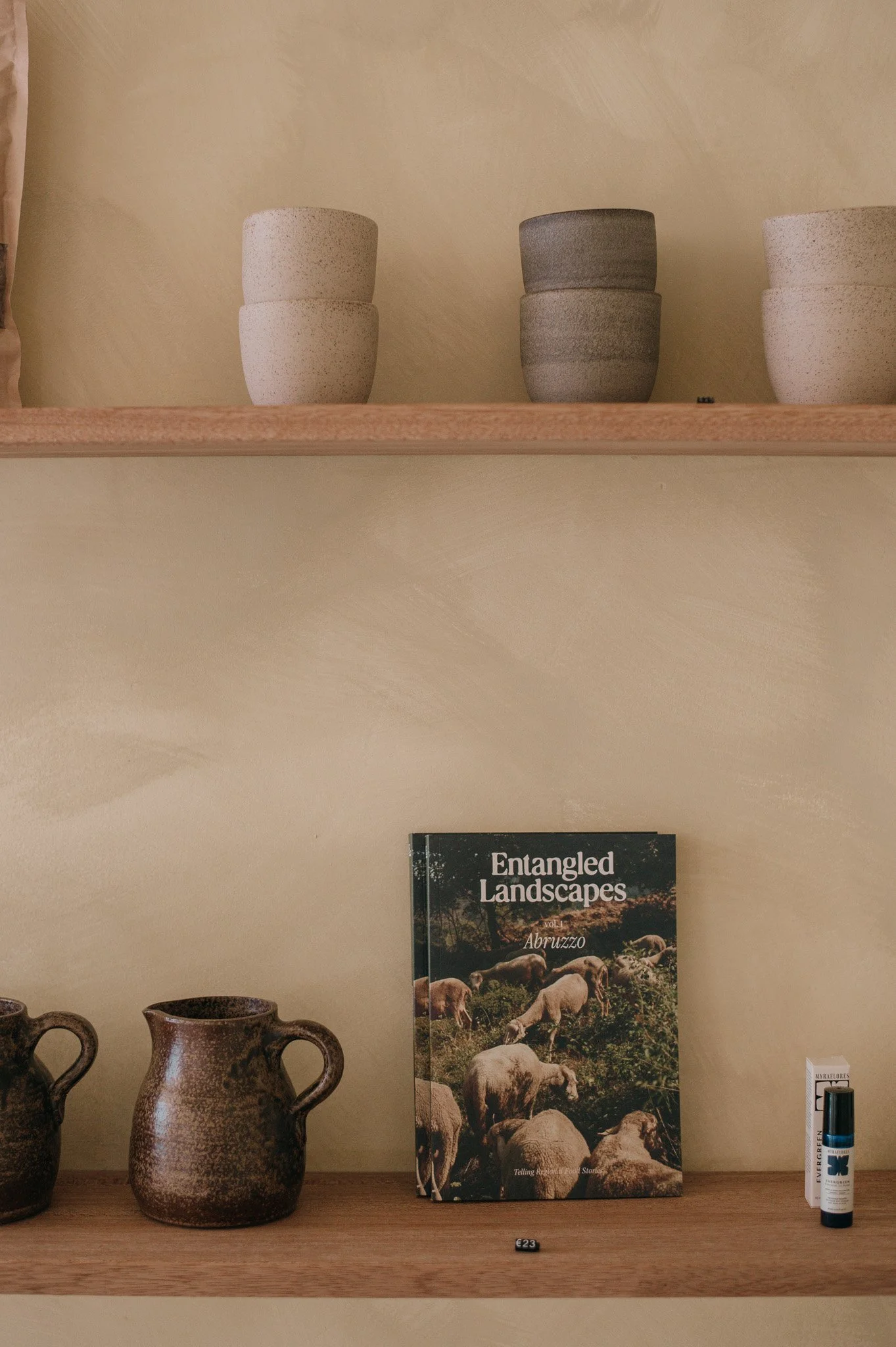 A wooden shelf with ceramic cups on top, a book titled "Entangled Landscapes" by Abruzzo, two brown ceramic pitchers, and a small bottle of essence on the bottom shelf.