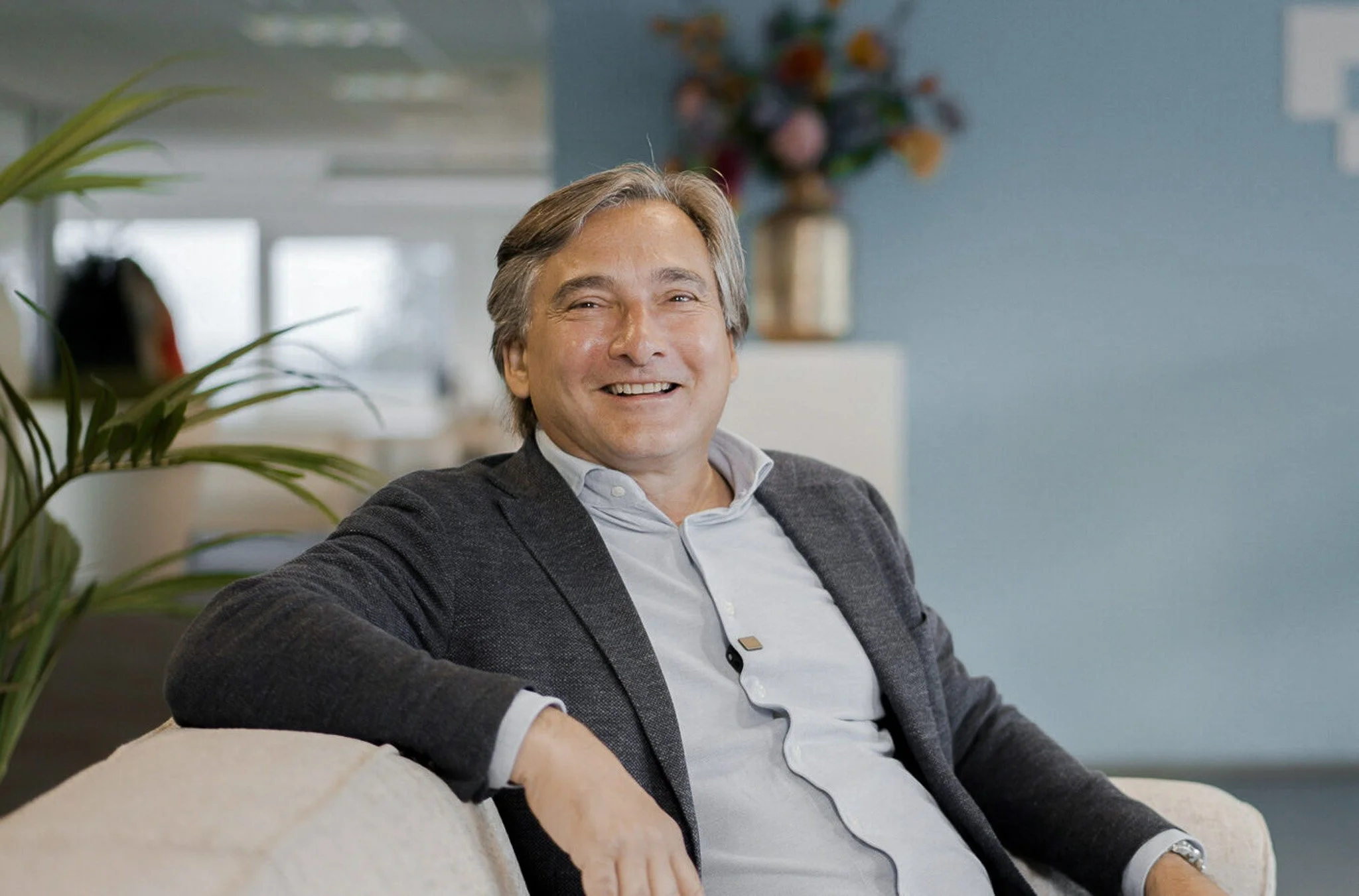 A middle-aged man with gray hair smiles while sitting on a beige sofa in a modern office environment background featuring a potted plant, a blue wall, a vase with flowers, and large windows.