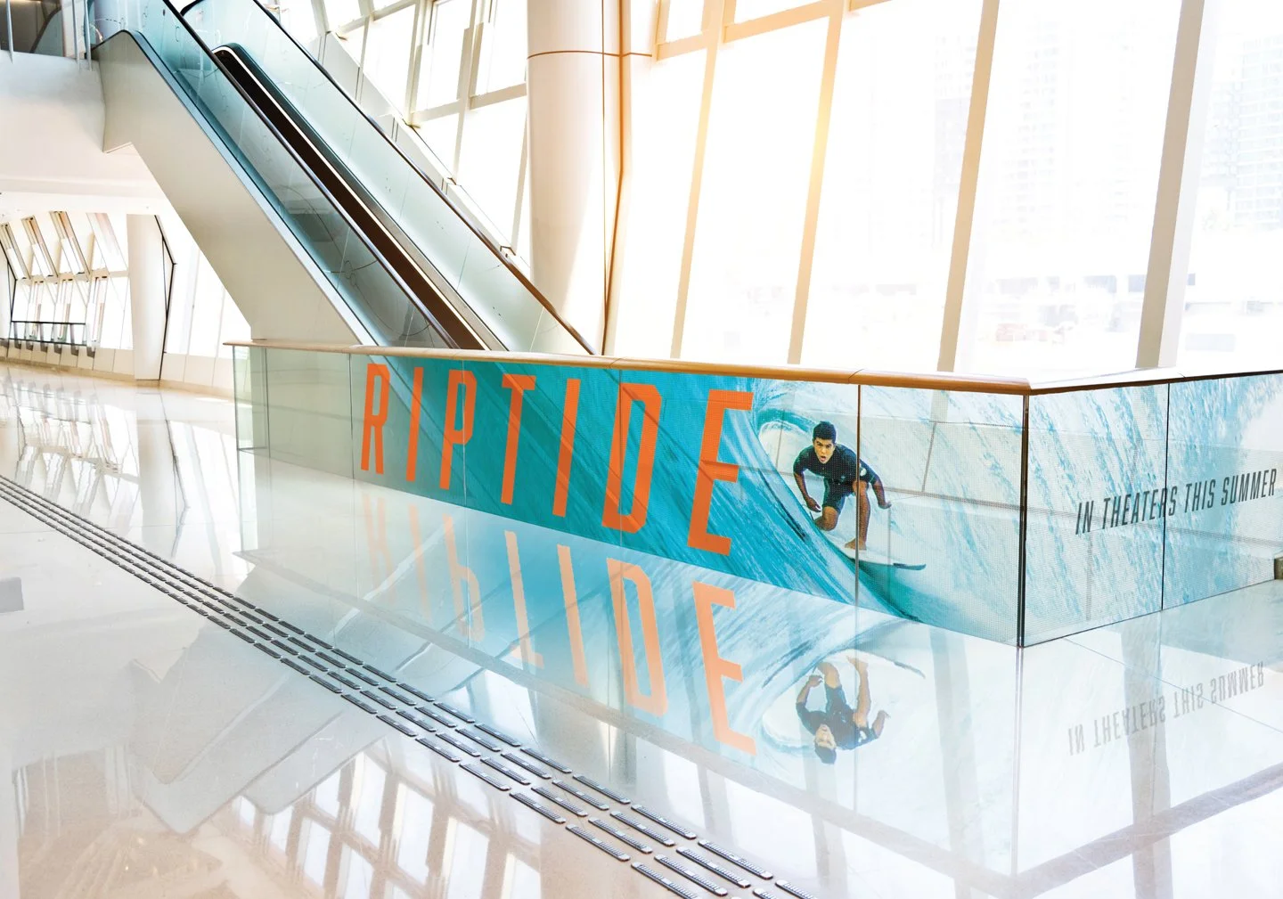 Interior of a modern building with large windows, an escalator, and LED glass advertising the movie "Riptide" with an image of a surfer and the text "In theaters this summer."