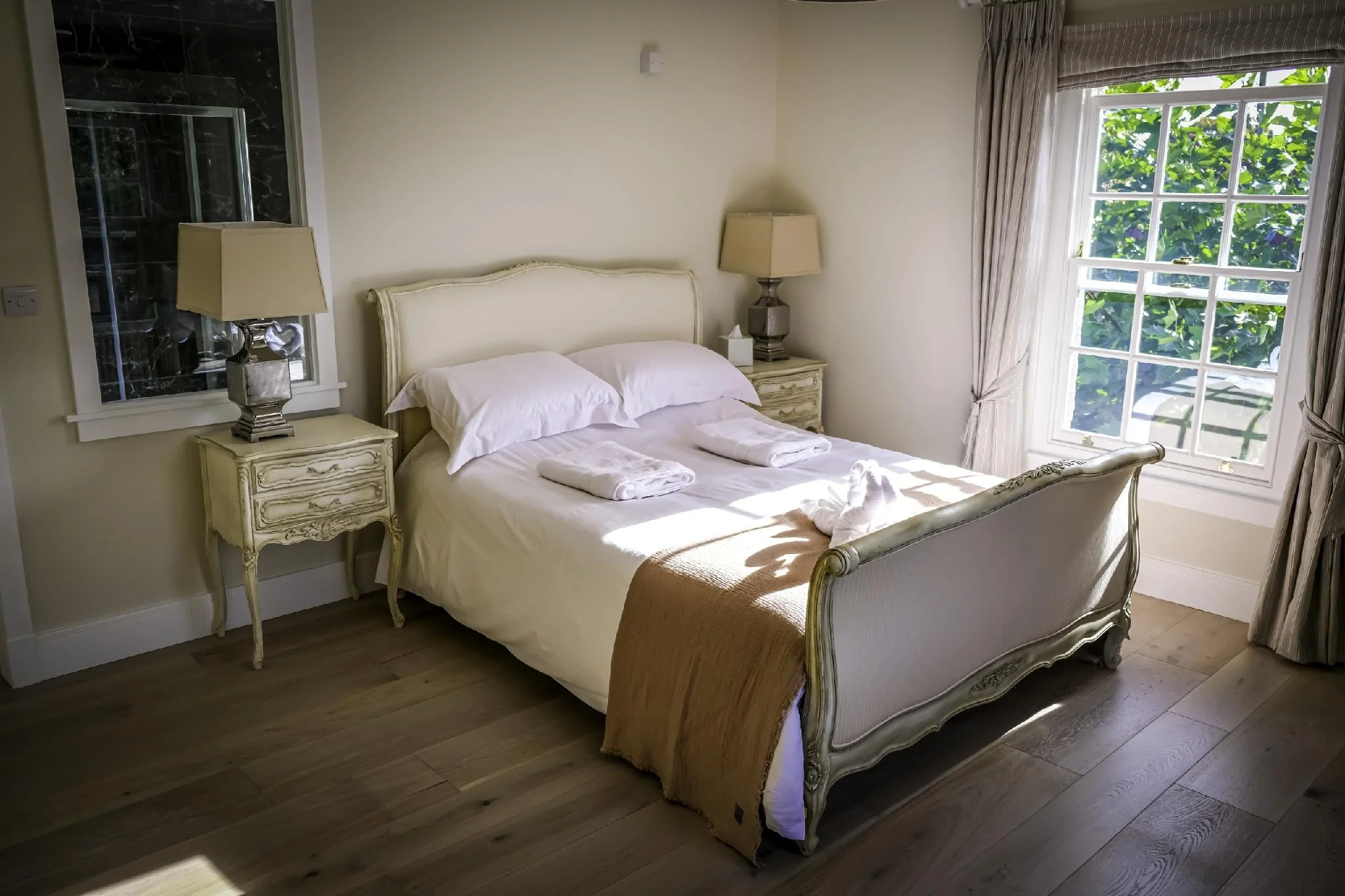 A well-made bed with white sheets, two pillows, and beige towels folded on top, flanked by two matching cream-colored nightstands with table lamps, in a bedroom with a large window showing green foliage outside, and light-colored hardwood floors.