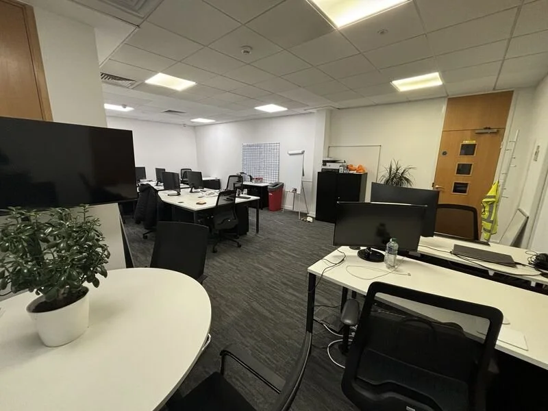 Office workspace with multiple desks, chairs, and monitors, a whiteboard, and a potted plant, illuminated by fluorescent ceiling lights.