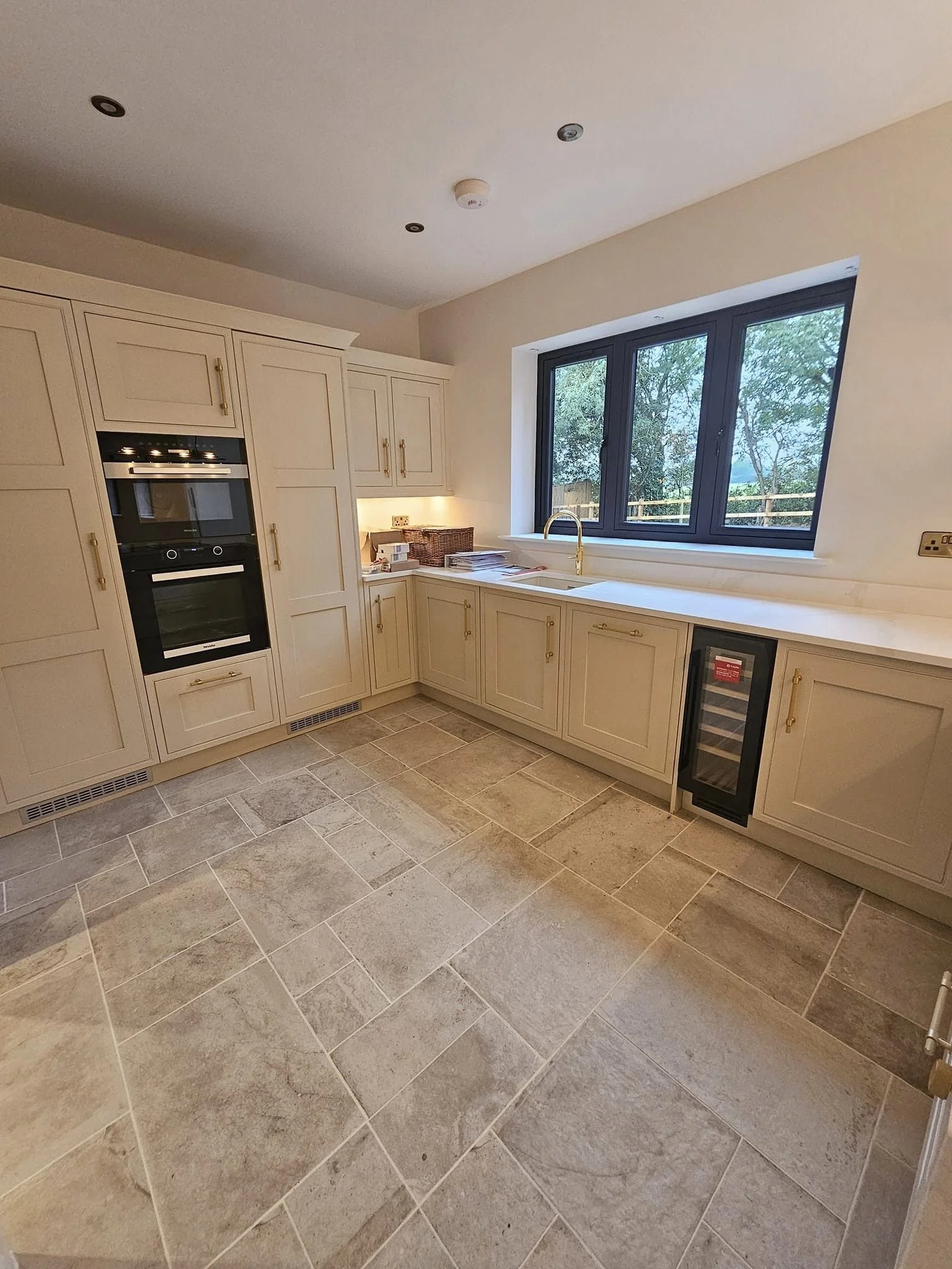 A modern kitchen with cream-colored cabinets, beige tiled flooring, a large window with a view of trees, and built-in appliances including a double oven and wine cooler.