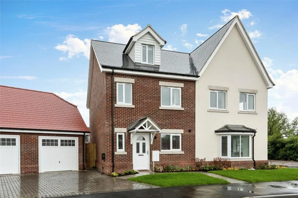 A two-story house with a brick facade on one side and an off-white exterior on the other, featuring a gable roof, a small covered porch, and several windows, with a separate garage to the left and a green lawn in front.