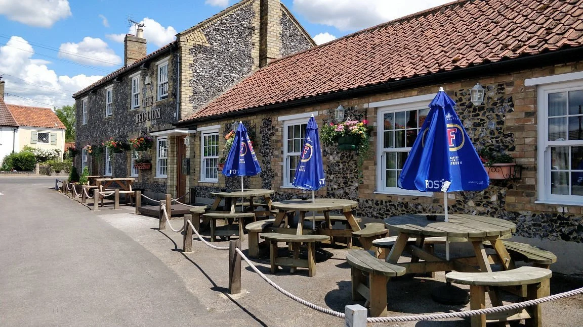 Outdoor patio with wooden picnic tables, blue umbrellas, and hanging flower baskets in front of a stone and brick building titled 'The Red Lion' on a sunny day.