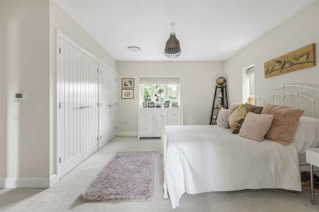 A minimalist bedroom with a white bed, multiple pillows, a white dresser, a ladder shelf, and a small window with greenery outside.