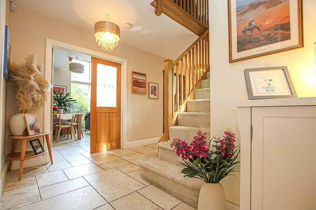Entrance hall with tiled floor, wooden staircase, beige carpet, white cabinet with framed picture, pink flowering plant, and a view into the dining area with natural light and green plants.