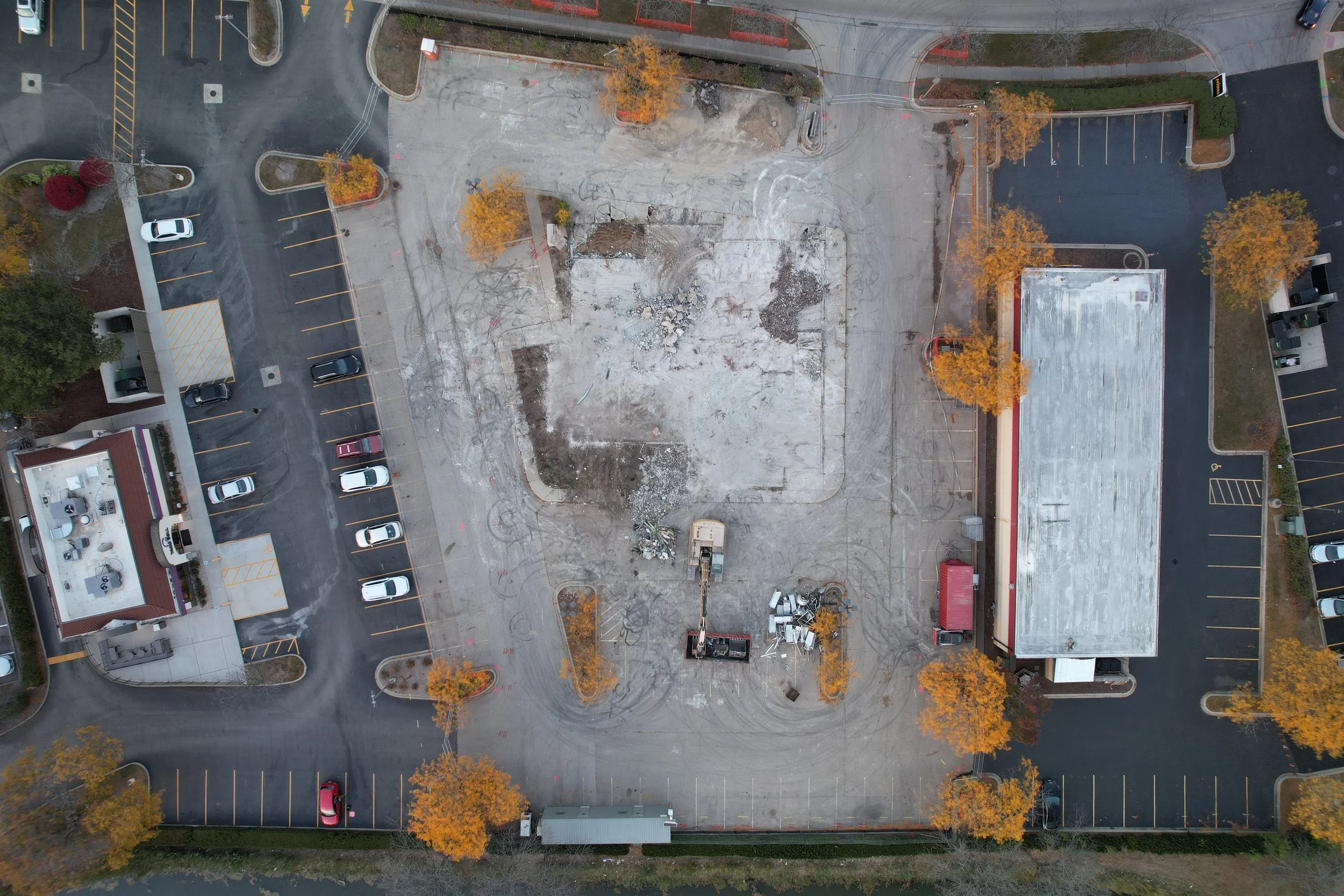 Aerial view of a building demolition site with construction equipment, surrounded by parking lots and trees with fall foliage.