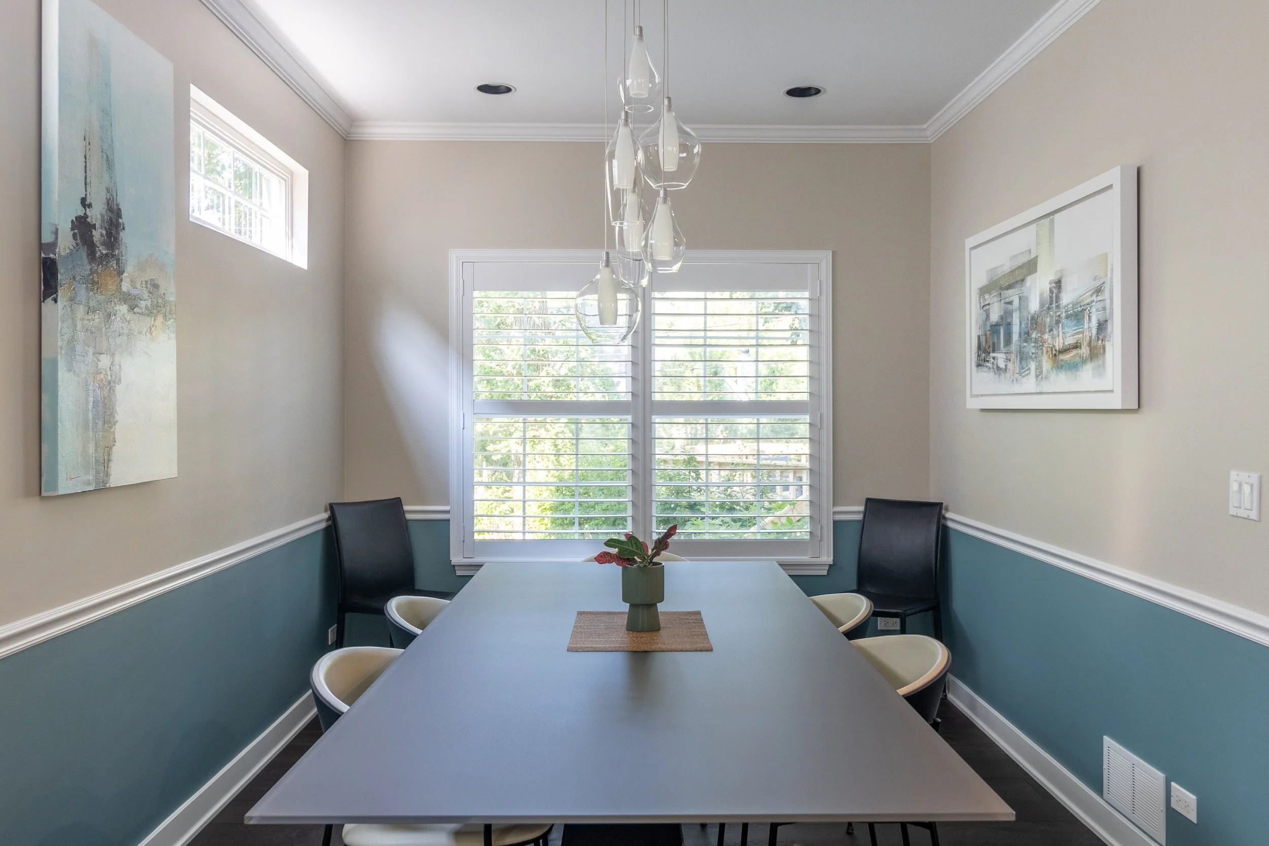 Dining room with a rectangular table, six chairs, a vase with a plant in the center, two paintings on the walls, a large window with blinds, and a hanging light fixture.