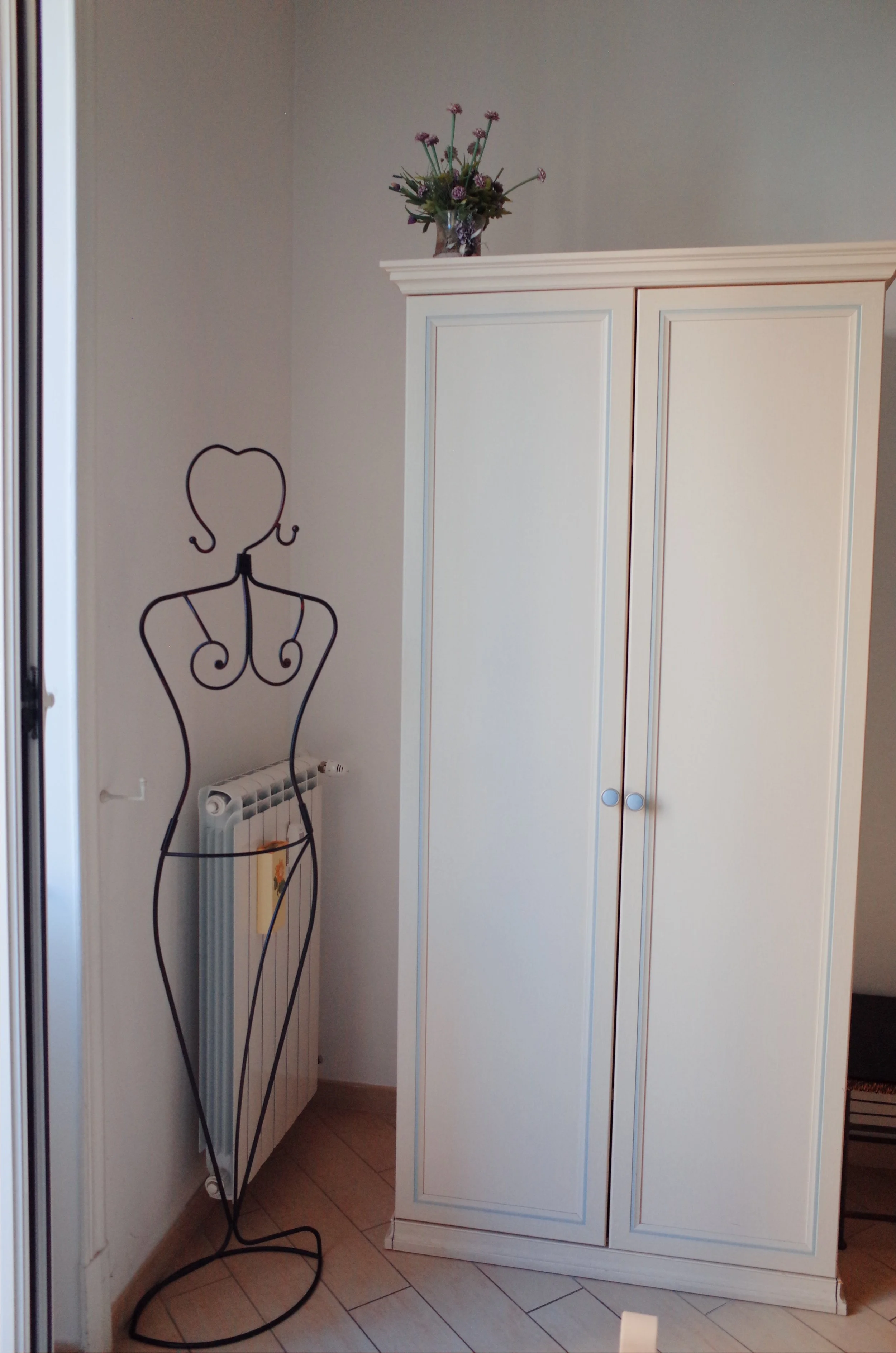 White wardrobe with decorative molding and a floral vase on top, located in a room corner. A black wire dress form stands beside a radiator below the wardrobe. The room has light-colored flooring and walls.