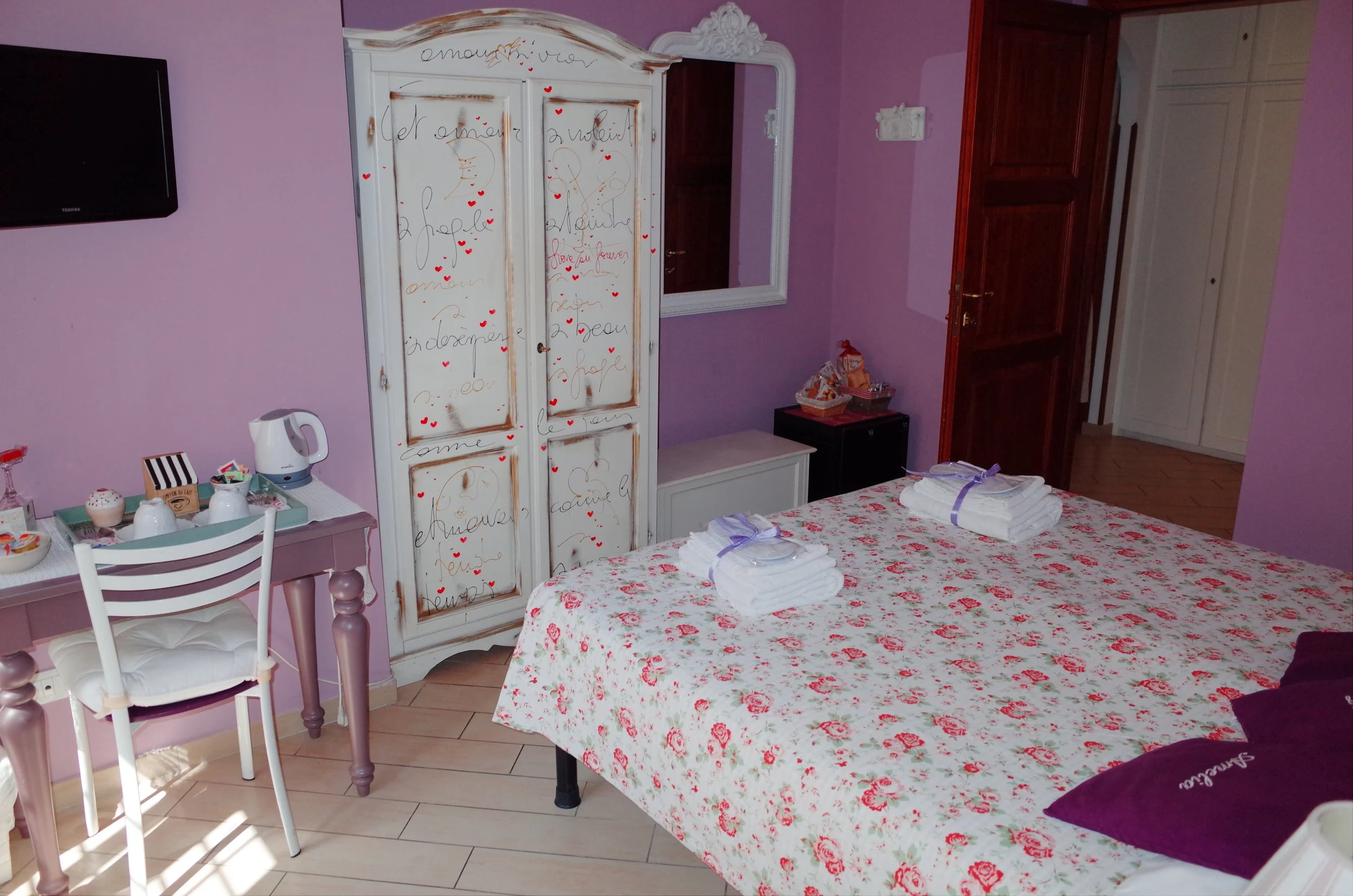 A cozy bedroom with pastel purple walls, a bed with a floral bedspread, and neatly folded white towels on top. There is a white wardrobe decorated with colorful handwritten words and small red hearts, a large white-framed mirror, and a small black ta