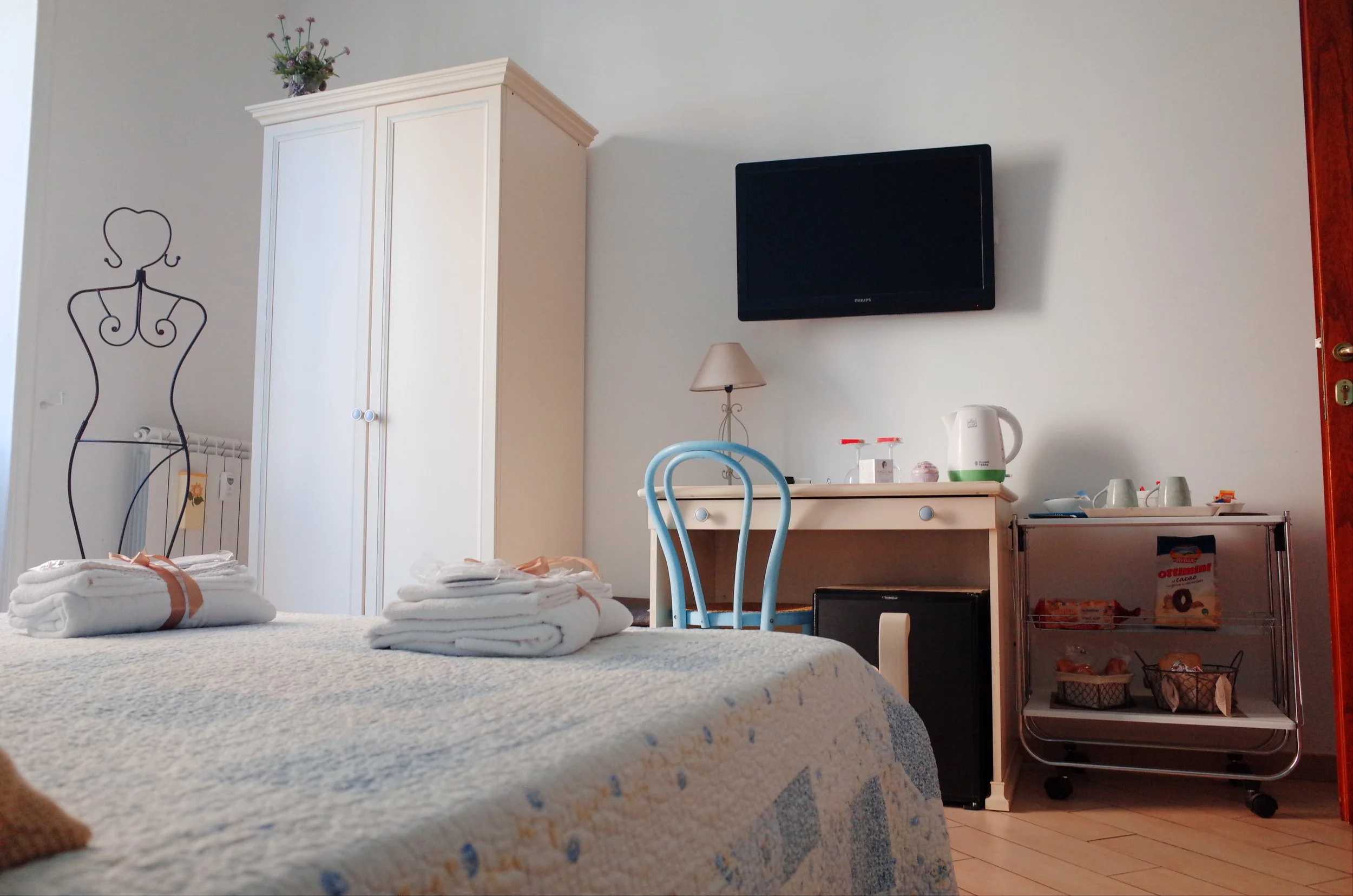 A hotel room with a bed in the foreground covered with a light-colored blanket and stacked towels. In the background, a white wardrobe, a wall-mounted TV, a small table with a lamp, a kettle, and cups. There is a decorative silhouette of a woman on t