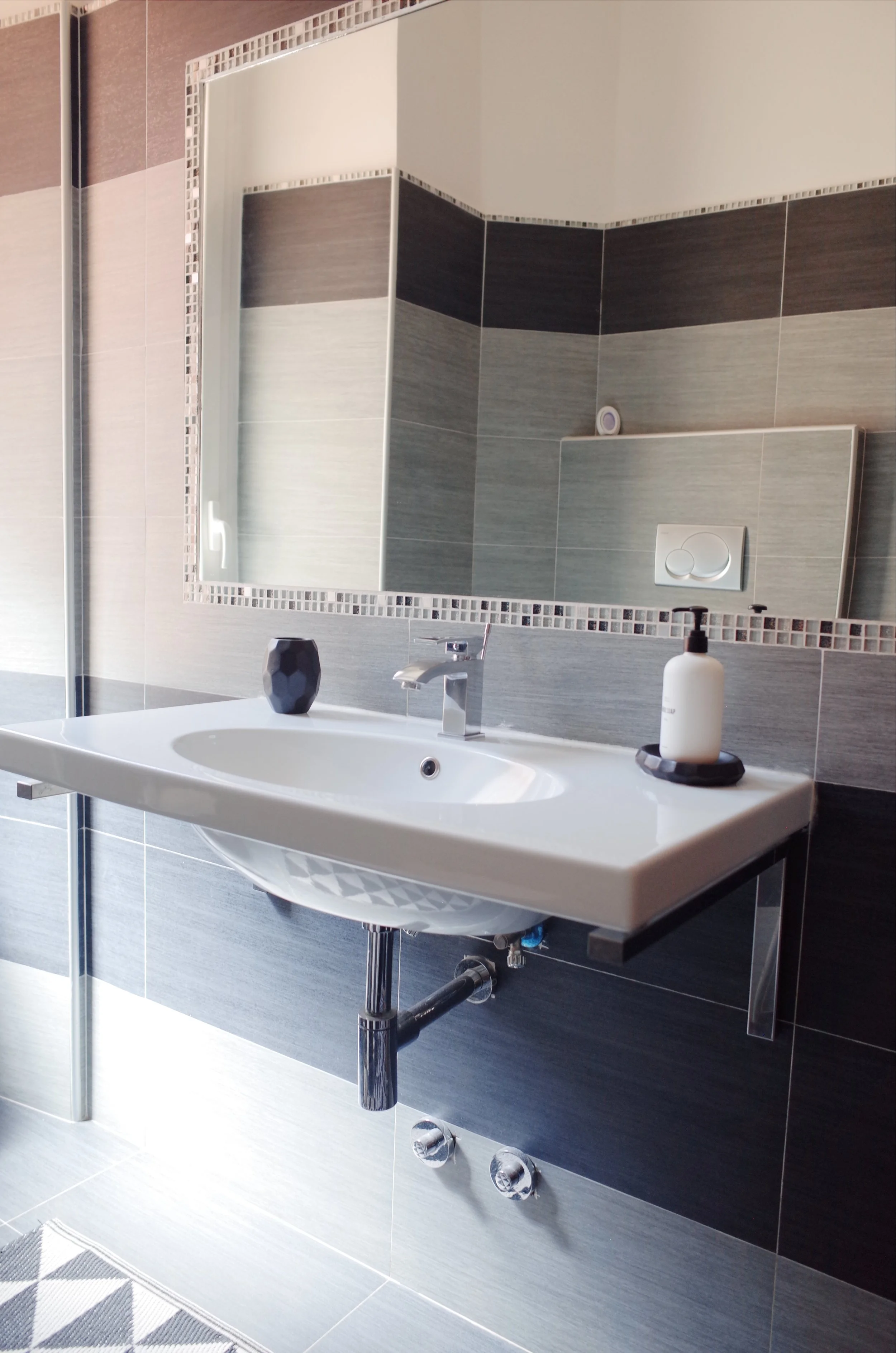 Modern bathroom with a white wall-mounted sink, a large mirror, gray tiles with darker and lighter shades, a soap dispenser, a small black decorative container, and a black and gray patterned floor mat.