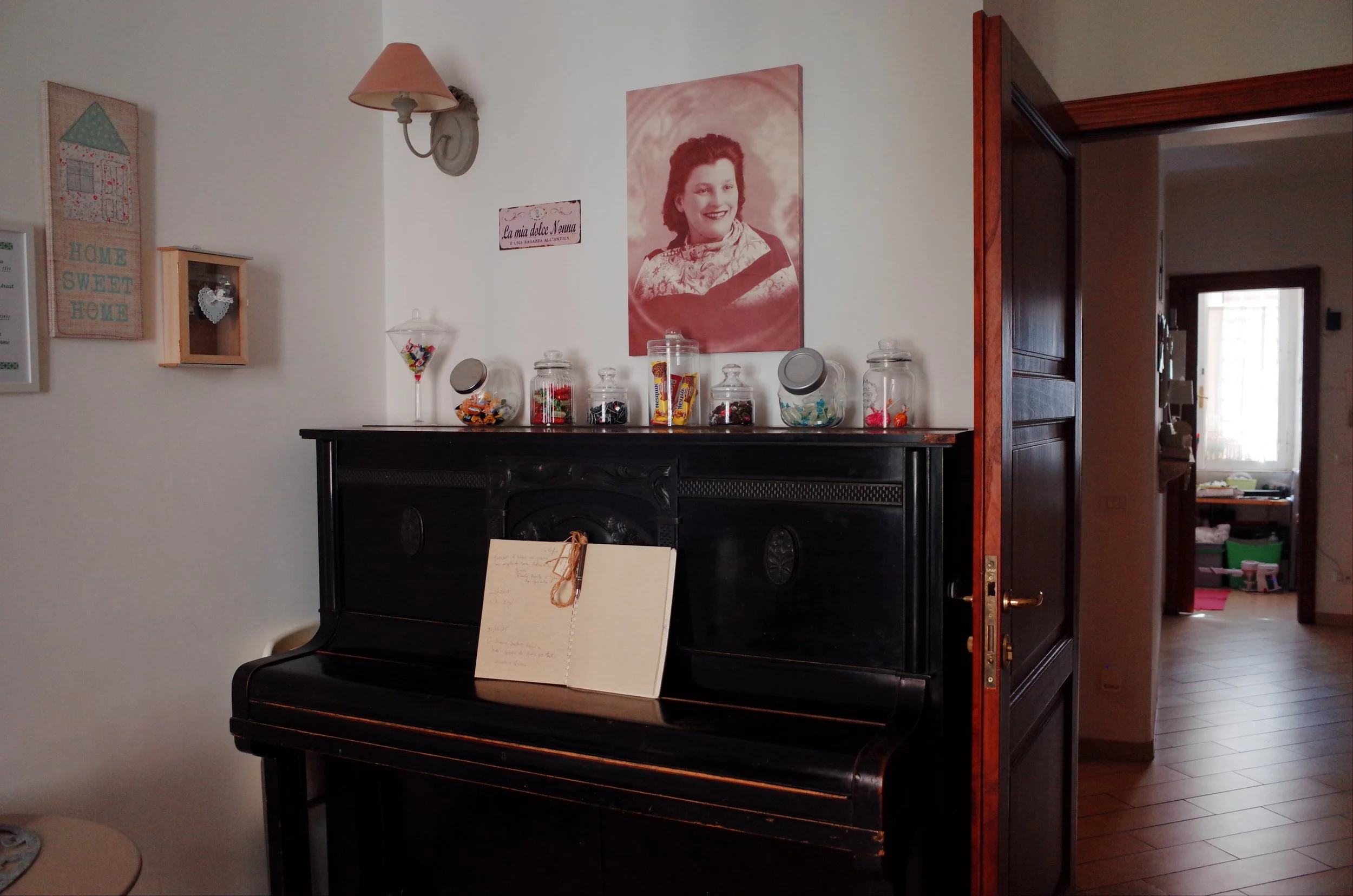 A black upright piano with a sheet of music on the music stand in a room with white walls and a wooden floor. On top of the piano are five glass jars filled with various candies and snacks. Behind the piano, on the wall, is a large black-and-white po