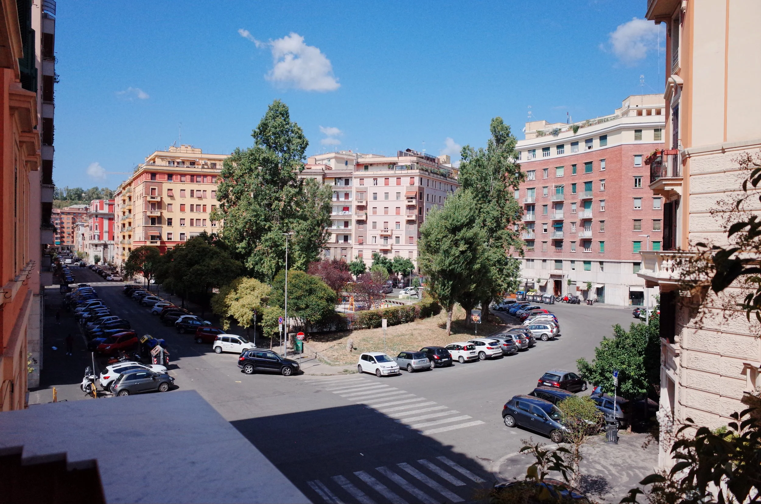 A city street view with several multi-story residential buildings, parked cars, trees, a blue sky with some clouds, and a crosswalk at the street corner.