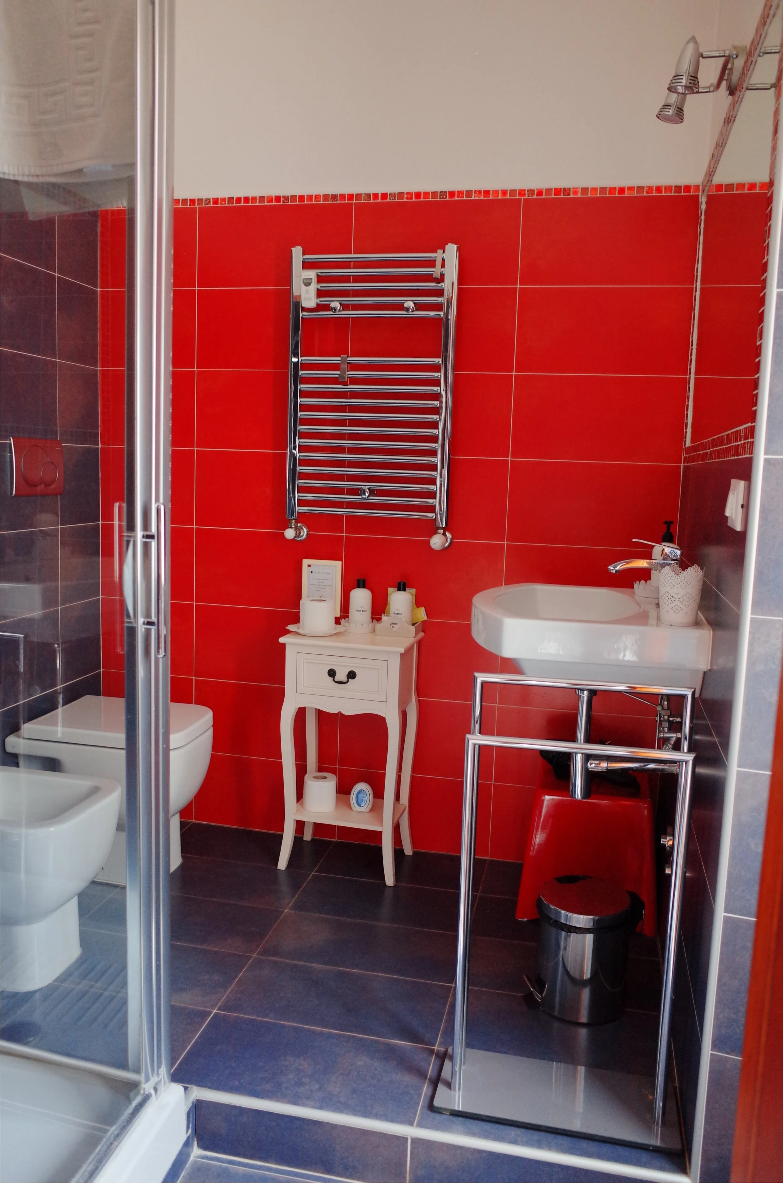 A modern bathroom with red tile walls, a small white side table holding toiletries, a white sink with a soap dispenser, a red trash bin, a stainless steel heated towel rail, and a shower area with a glass door.