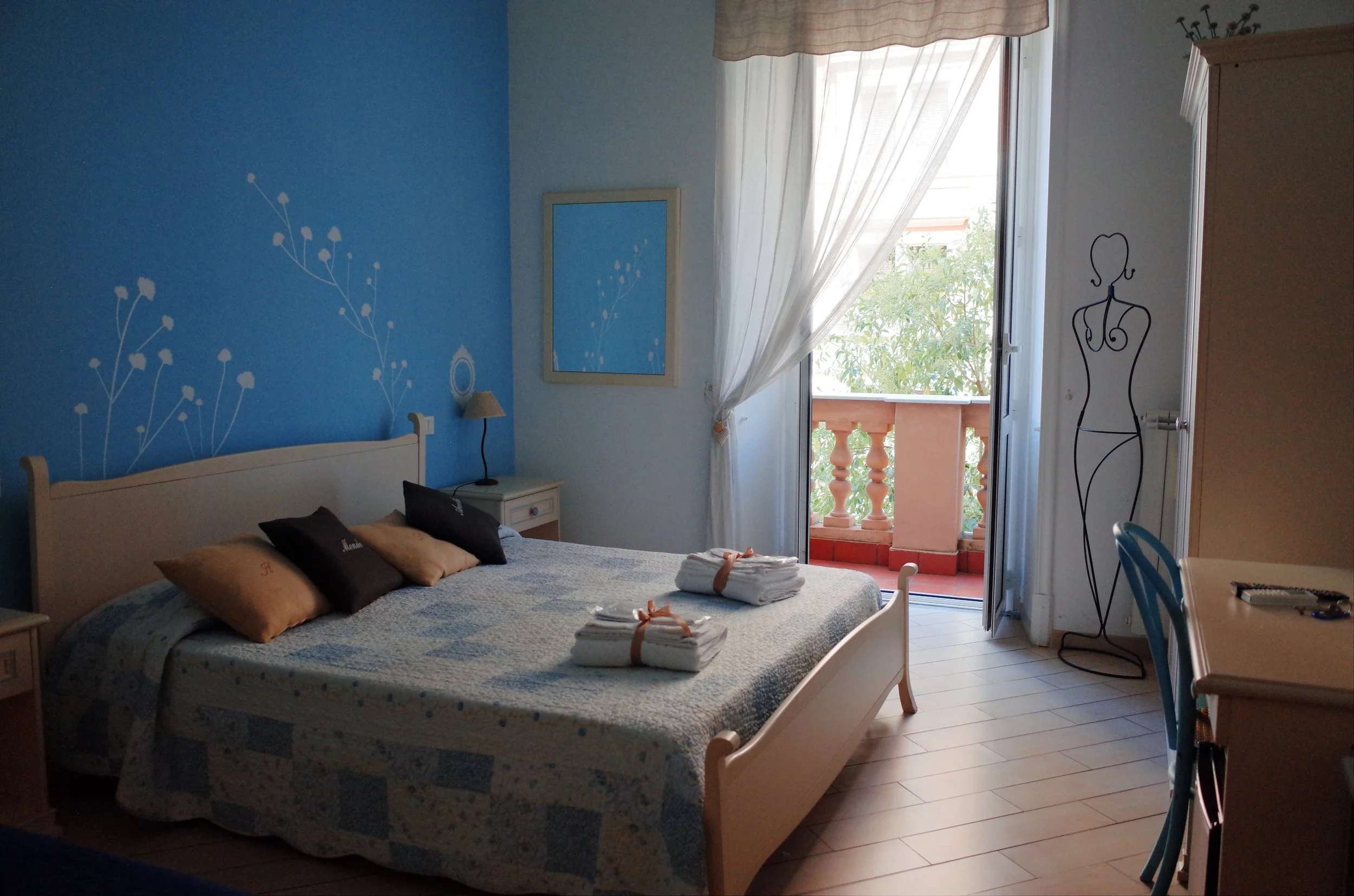 A bedroom with a blue accent wall, a bed with brown and black pillows, stacked towels on the bed, a nightstand with a lamp, a mirror on the wall, a mannequin outline near the door, and open glass doors leading to a balcony with trees outside.