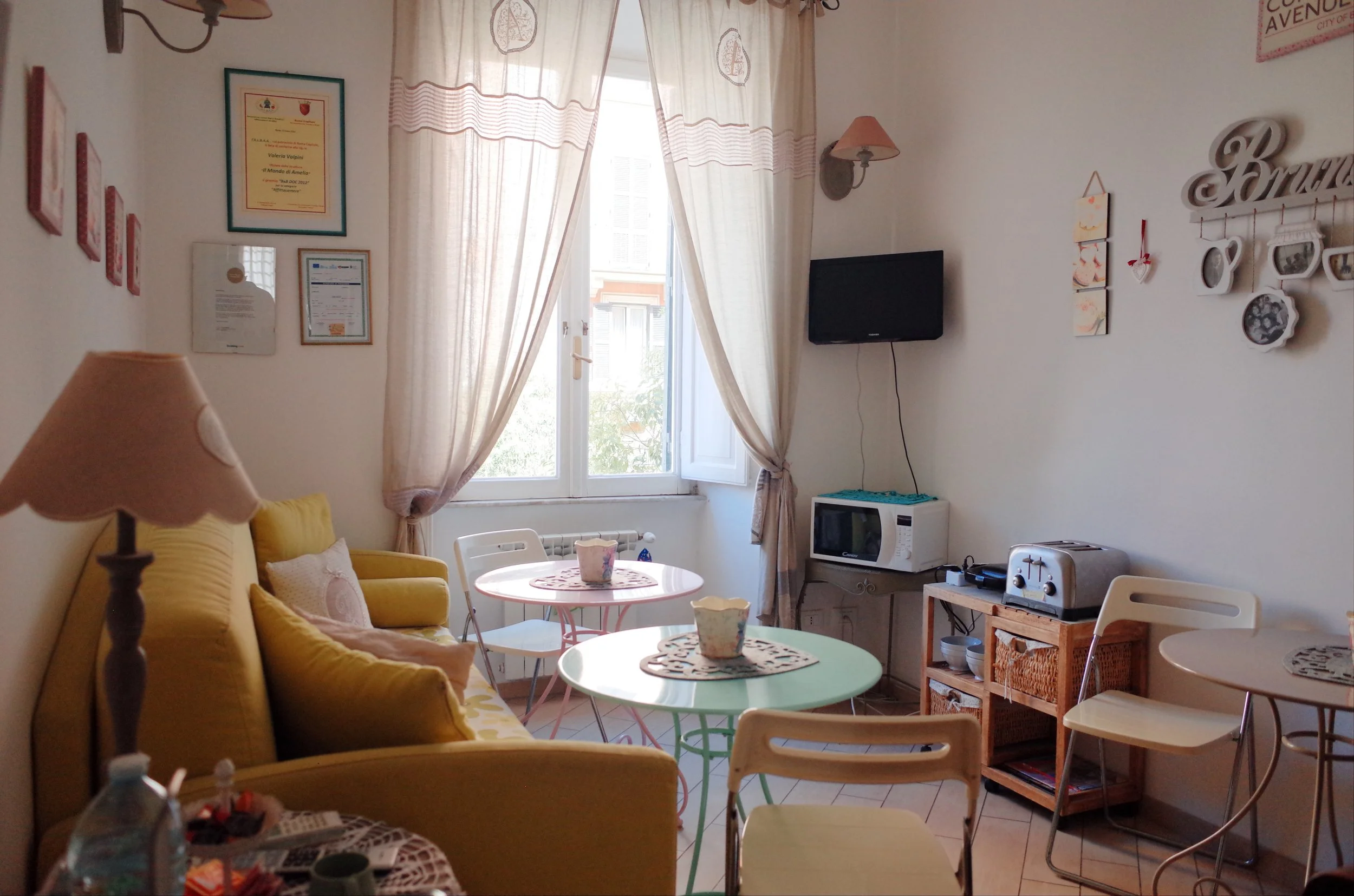 A cozy living room with yellow sofa, small round tables with floral cups, a lamp, a wall-mounted TV, wall decorations, a microwave, toaster, and various wall plaques and photographs near a window with beige curtains letting in natural light.