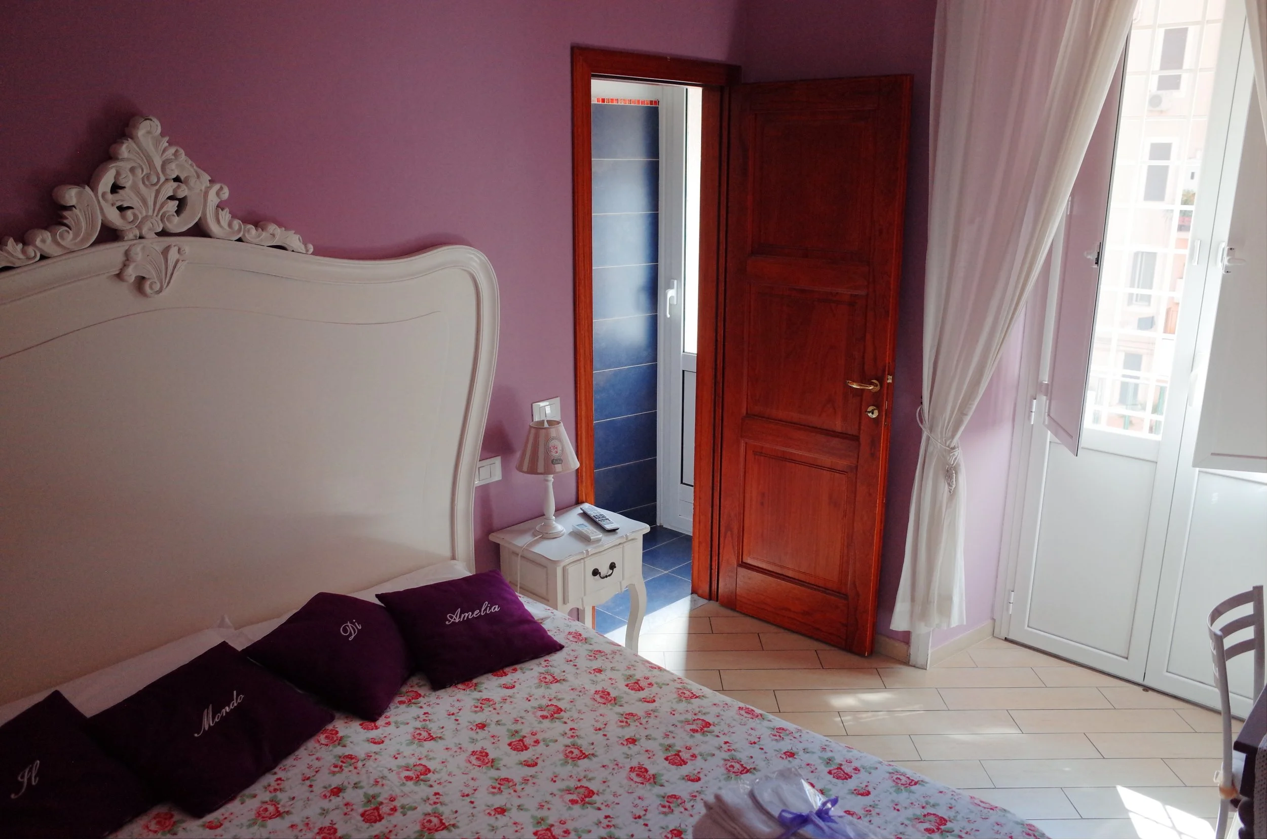 A bedroom with a white ornate headboard, purple pillows, and a floral bedspread. There is a white nightstand with a lamp and remote control. The room has purple walls, wooden trim, and a window with white curtains. An open door shows a balcony or ano
