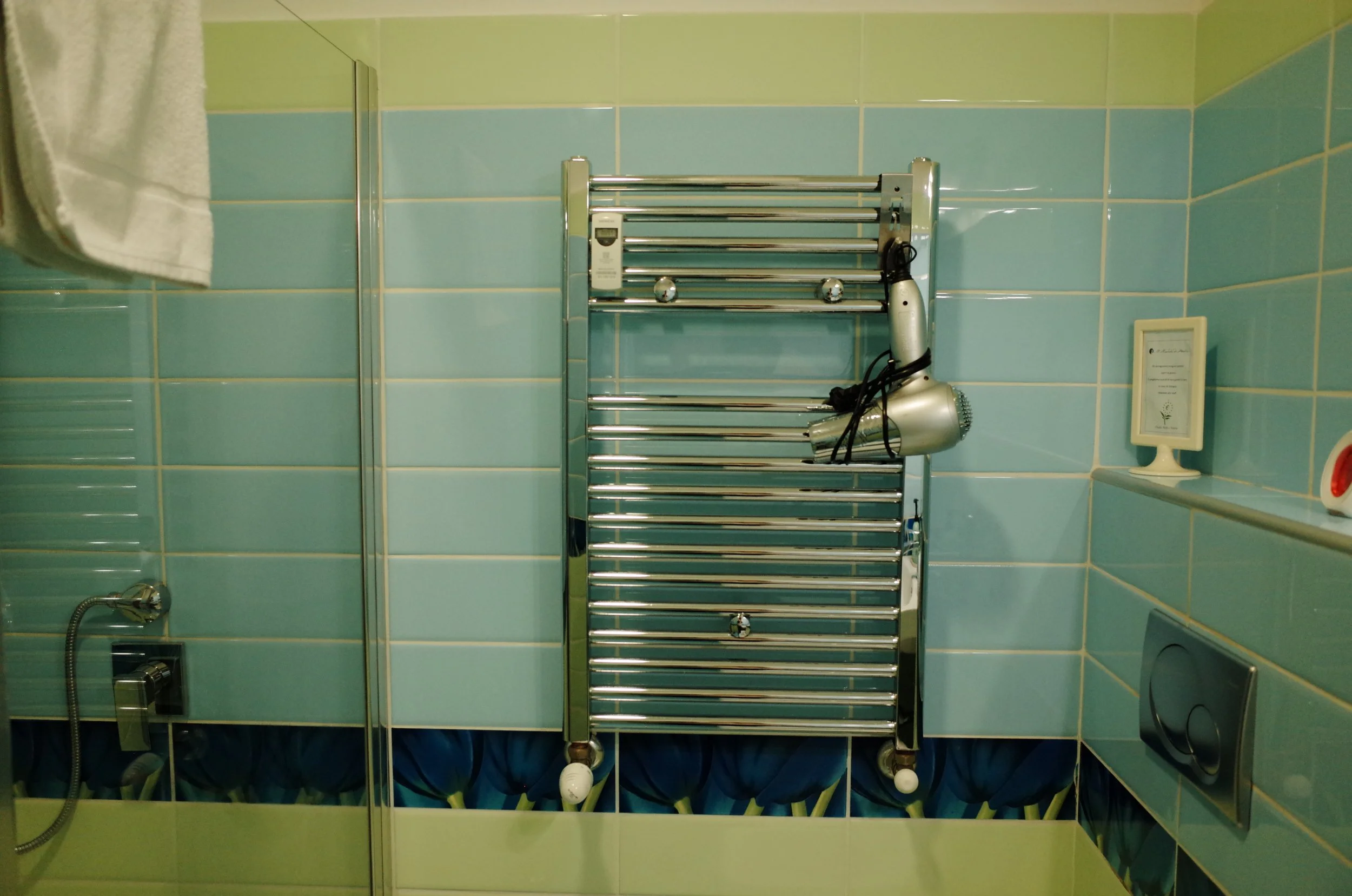 A bathroom with green and blue tiled walls, a heated towel rack with a hairdryer hanging on it, a framed certificate or document on the wall, and part of a shower head visible.