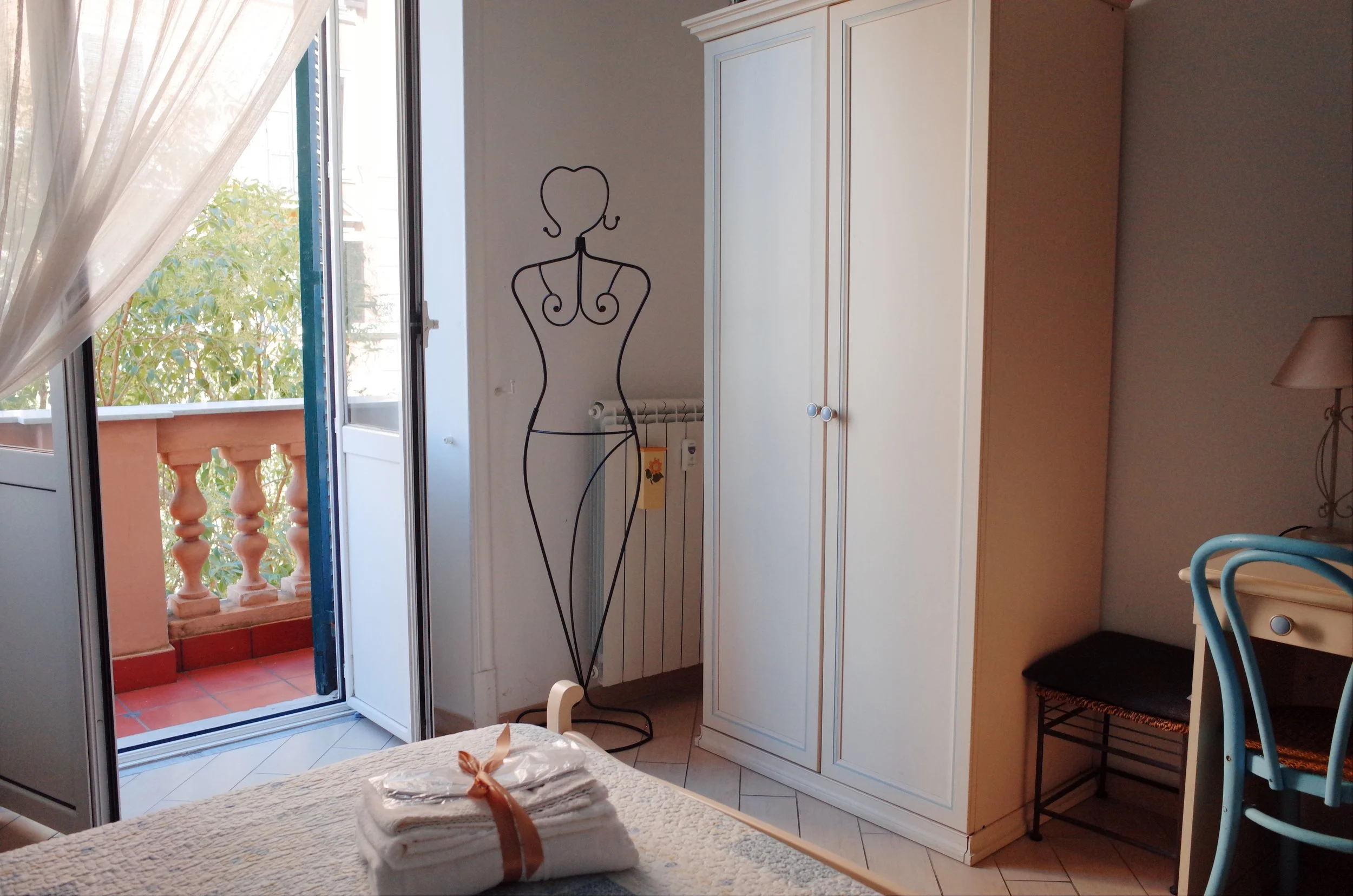 A room with a white wardrobe, a metal dress form, and an open door leading to a small balcony with a terracotta railing, some greenery outside, and a sheer curtain partially covering the door.