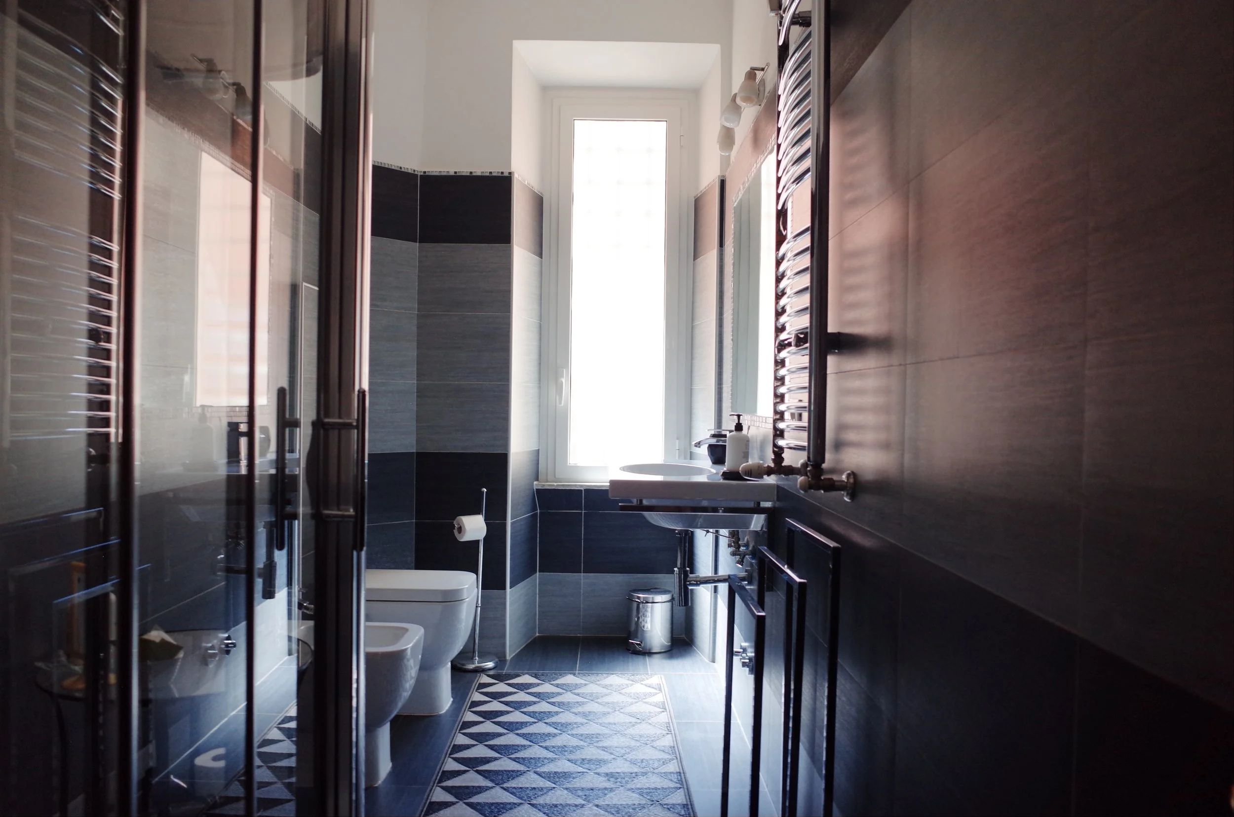 Modern bathroom with black and gray tiled walls, toilet, sink, and a window allowing natural light, with decorative black and white patterned floor tiles.