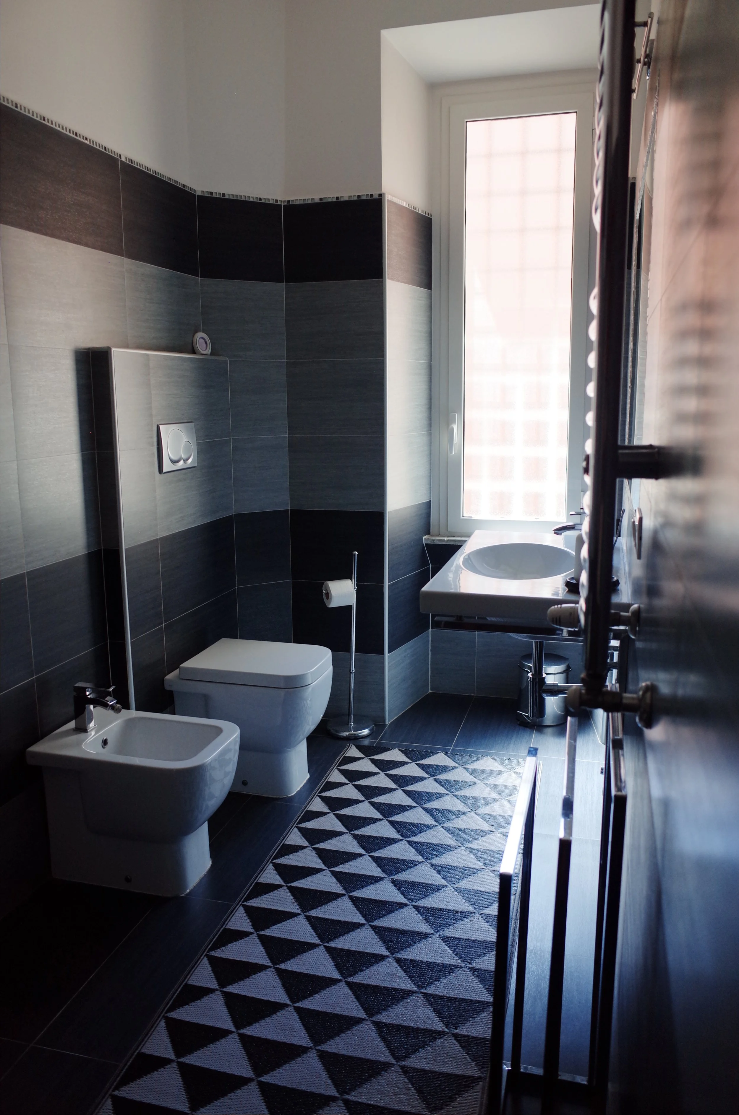 Modern bathroom with dark gray and light gray tiled walls, a window letting in natural light, a wall-mounted toilet, a bidet, a small sink, a geometric black and white rug, and metallic towel rails.