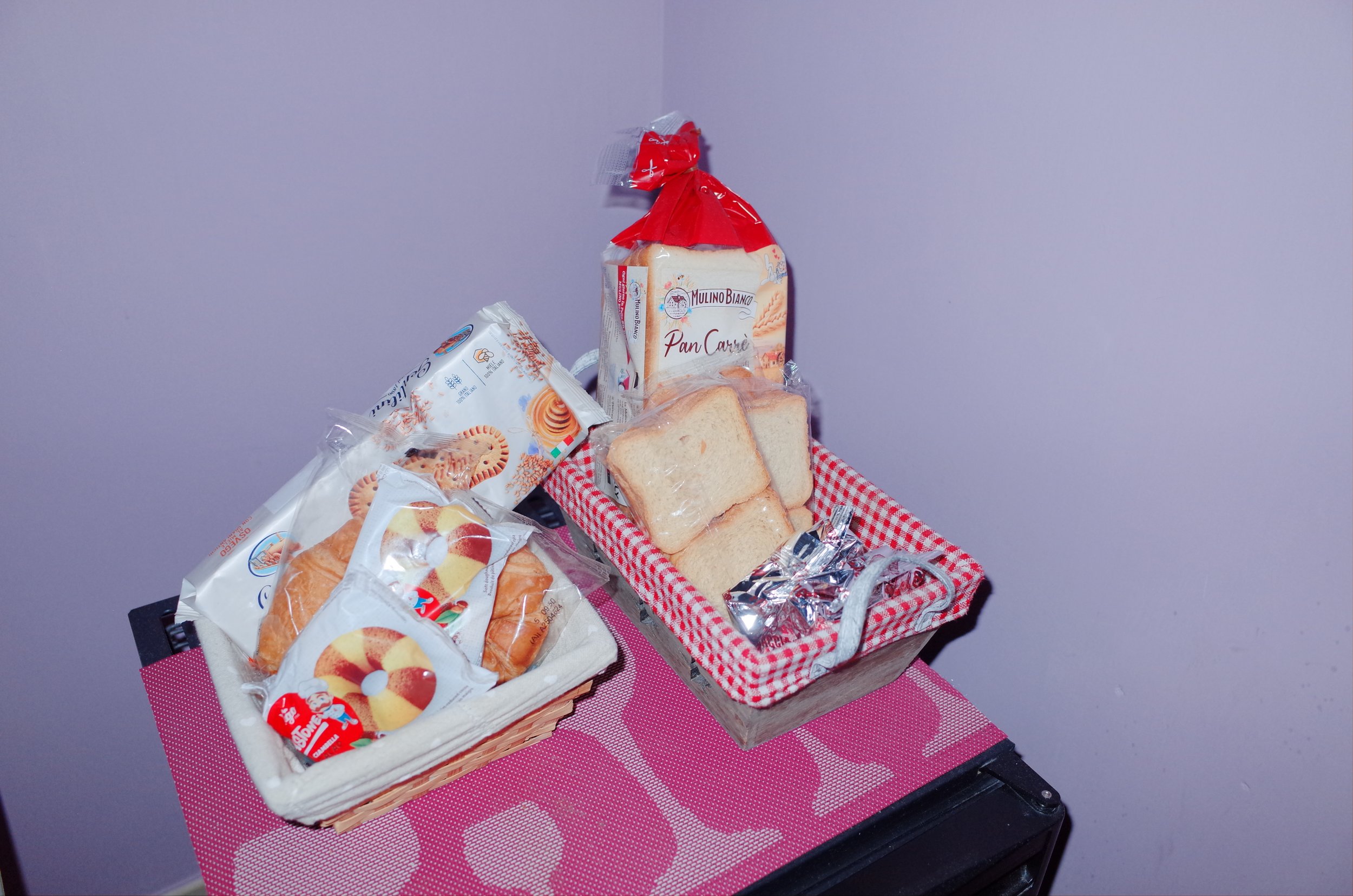 Assorted sliced bread and packaged cookies on a small table with a red checkered basket and a bag of bread against a purple wall background.