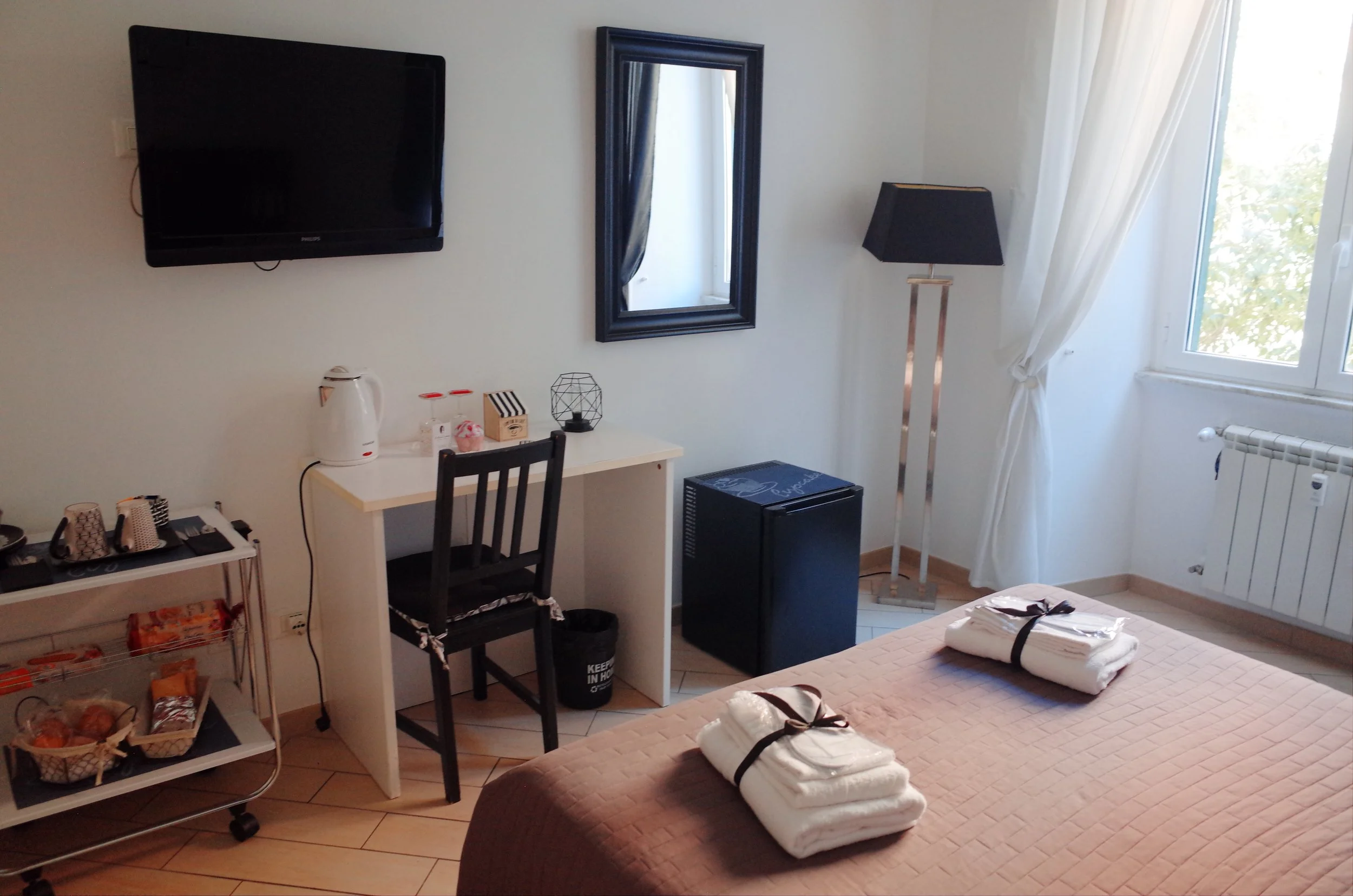 A cozy bedroom with a wall-mounted TV, a mirror, a standing lamp, and window with white curtains. There is a small desk with various items, a mini fridge, and a bed with folded towels on it.