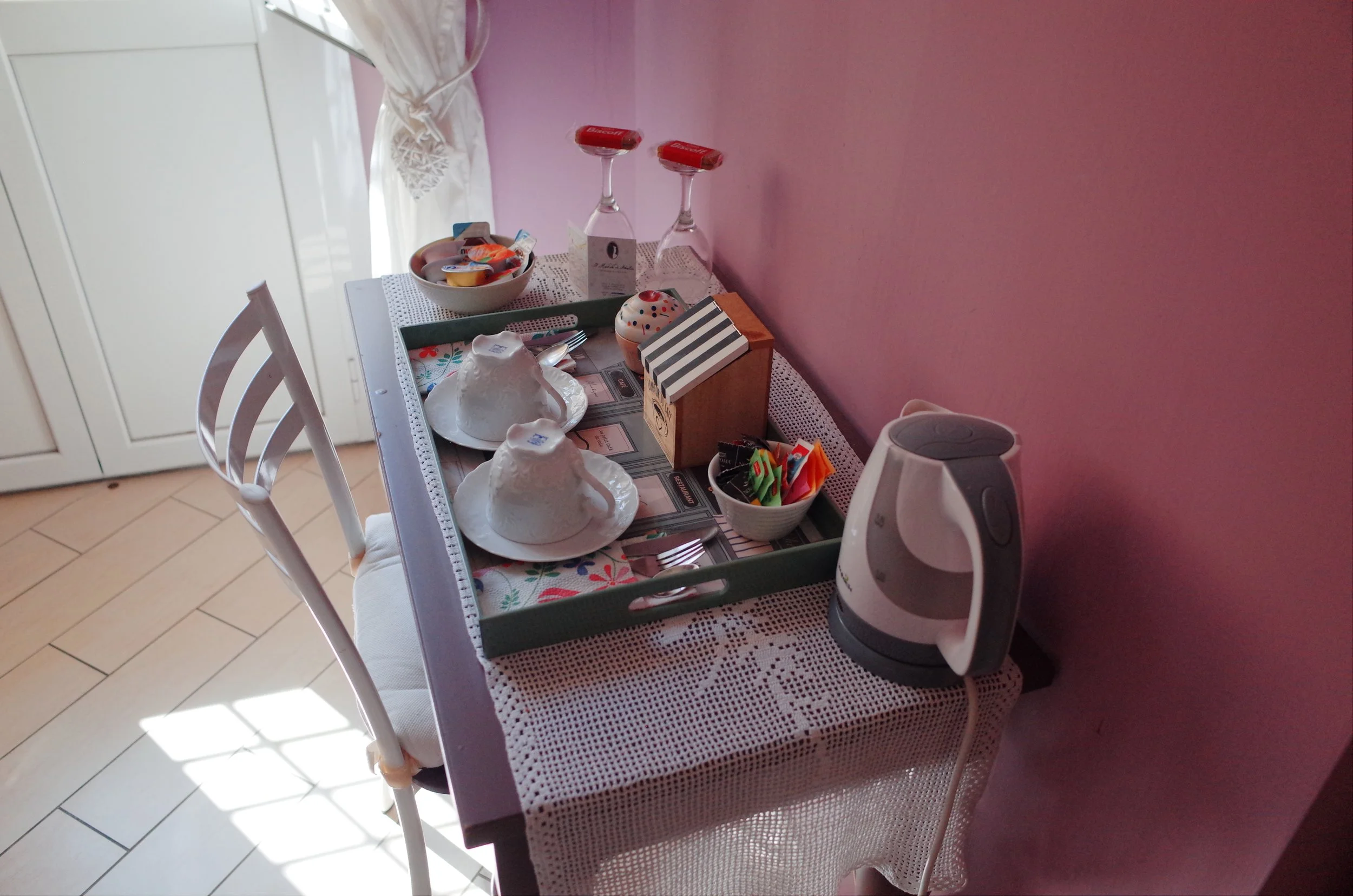 A dining table set for breakfast with two tea cups covered with a white lace napkin, a small bowl of yogurt with fruit, two upside-down wine glasses, a small wooden box, a bowl of tea bags, a toaster, and some utensils, against a pink wall.