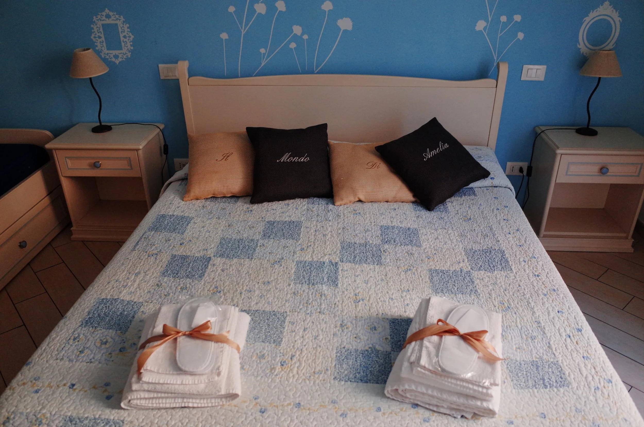 A bedroom with a blue wall, white furniture, and a quilted bedspread. Four pillows spell out 'Il Mondo Di Amelia' on the bed. Two bedside tables with lamps are on each side of the bed. In front of the bed, there are white wrapped gifts with peach rib