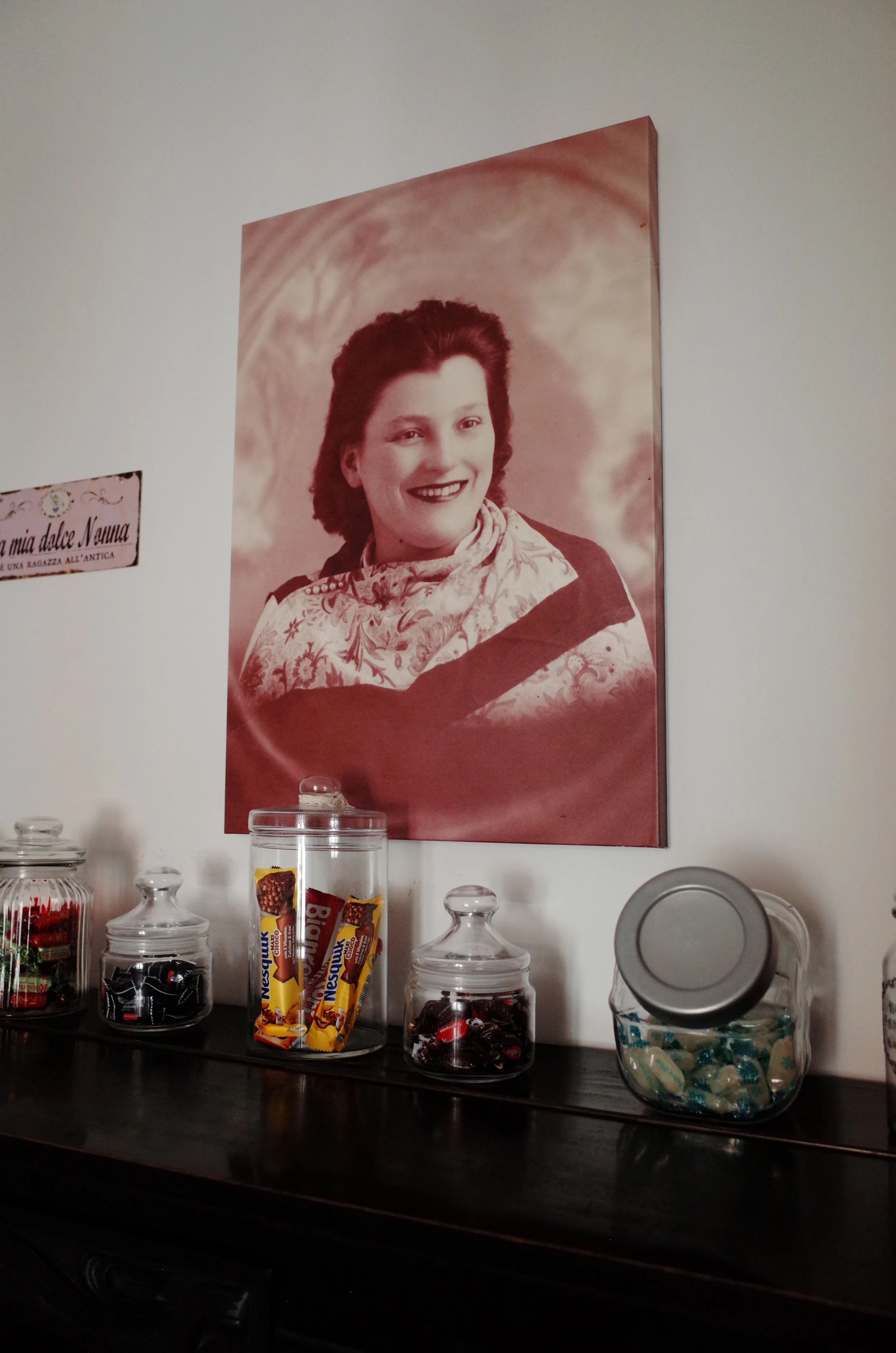 A wall with a large, vintage-style portrait of a woman with dark, wavy hair and a bright smile, hung above a wooden surface. The surface holds various glass jars and containers filled with candies and chocolates, some labeled 'Nestle Big Chocolates,'