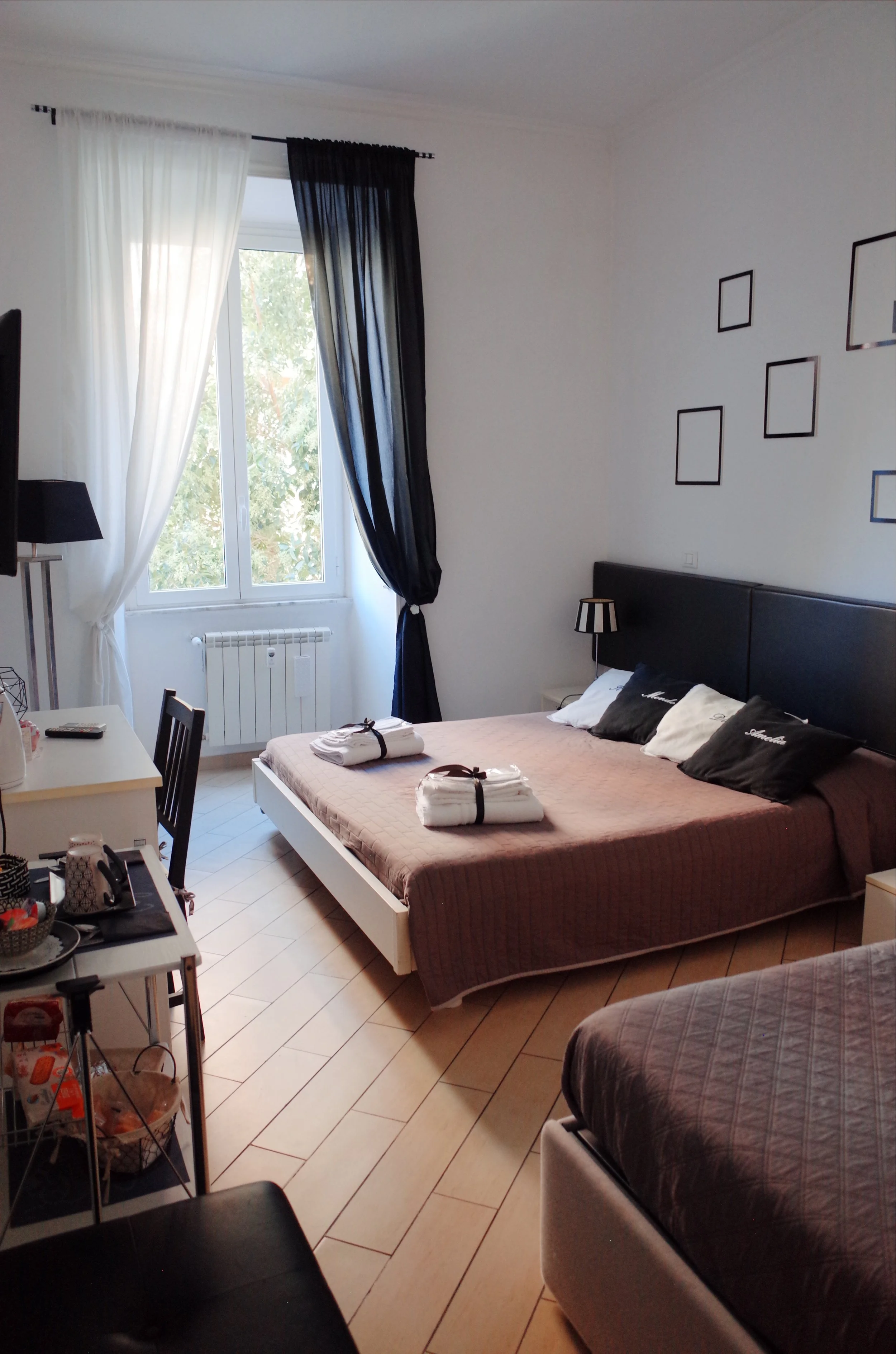 A bedroom with a bed, window with white and black curtains, a small bedside table with a lamp, a black armchair, and a dresser with various items, including neatly folded towels.