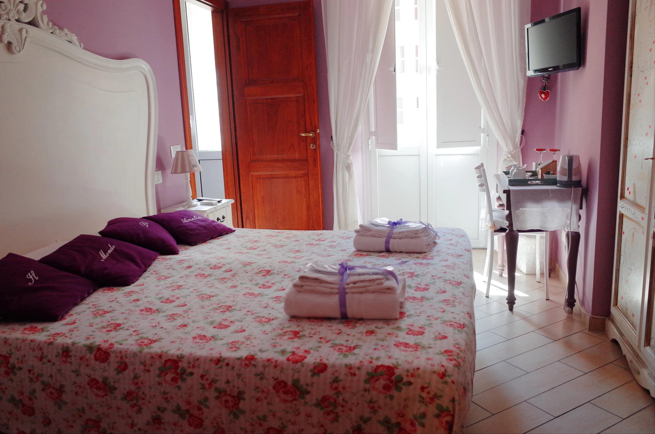 A cozy bedroom with a floral bedspread and purple pillows labeled with names, a wooden door, a window with white curtains, a flat-screen TV, a small white desk with a chair, and neatly folded towels on the bed.