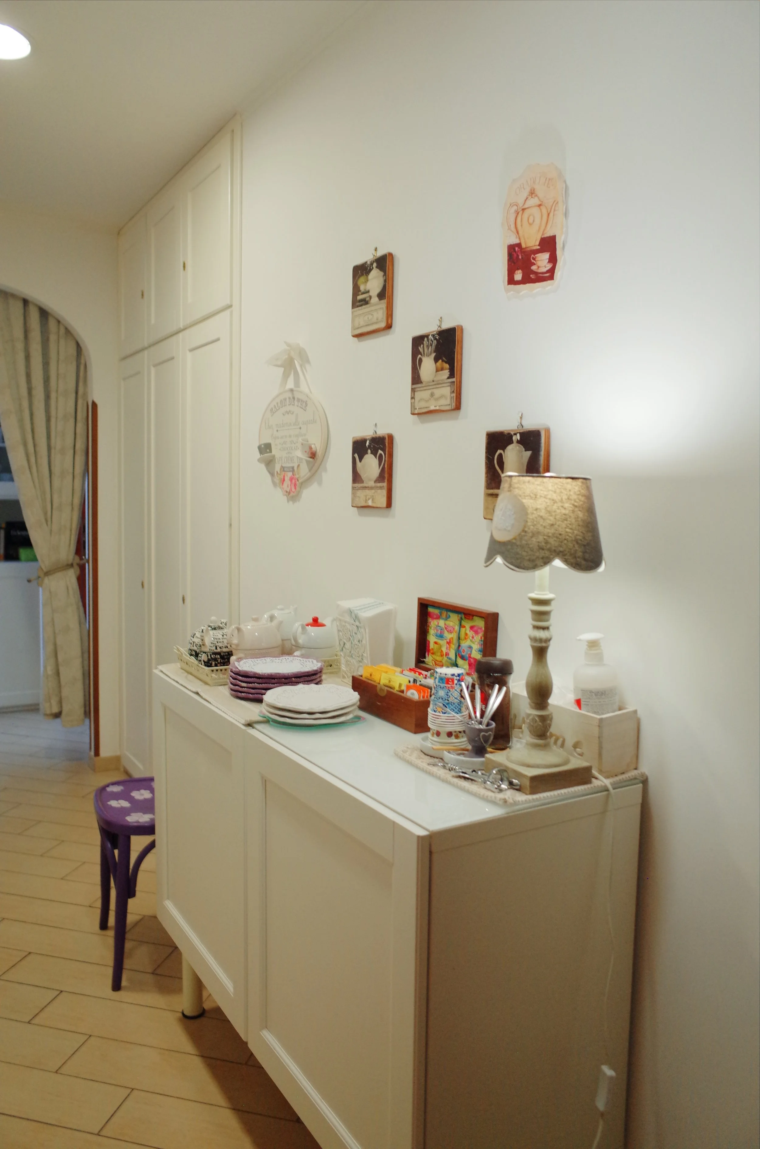 Interior view of a dining or breakfast area with a white sideboard, decorated with teapots, cups, and miscellaneous items. A purple stool with paw prints is in the foreground. The wall features hanging framed artwork and a round decorative wall hangi