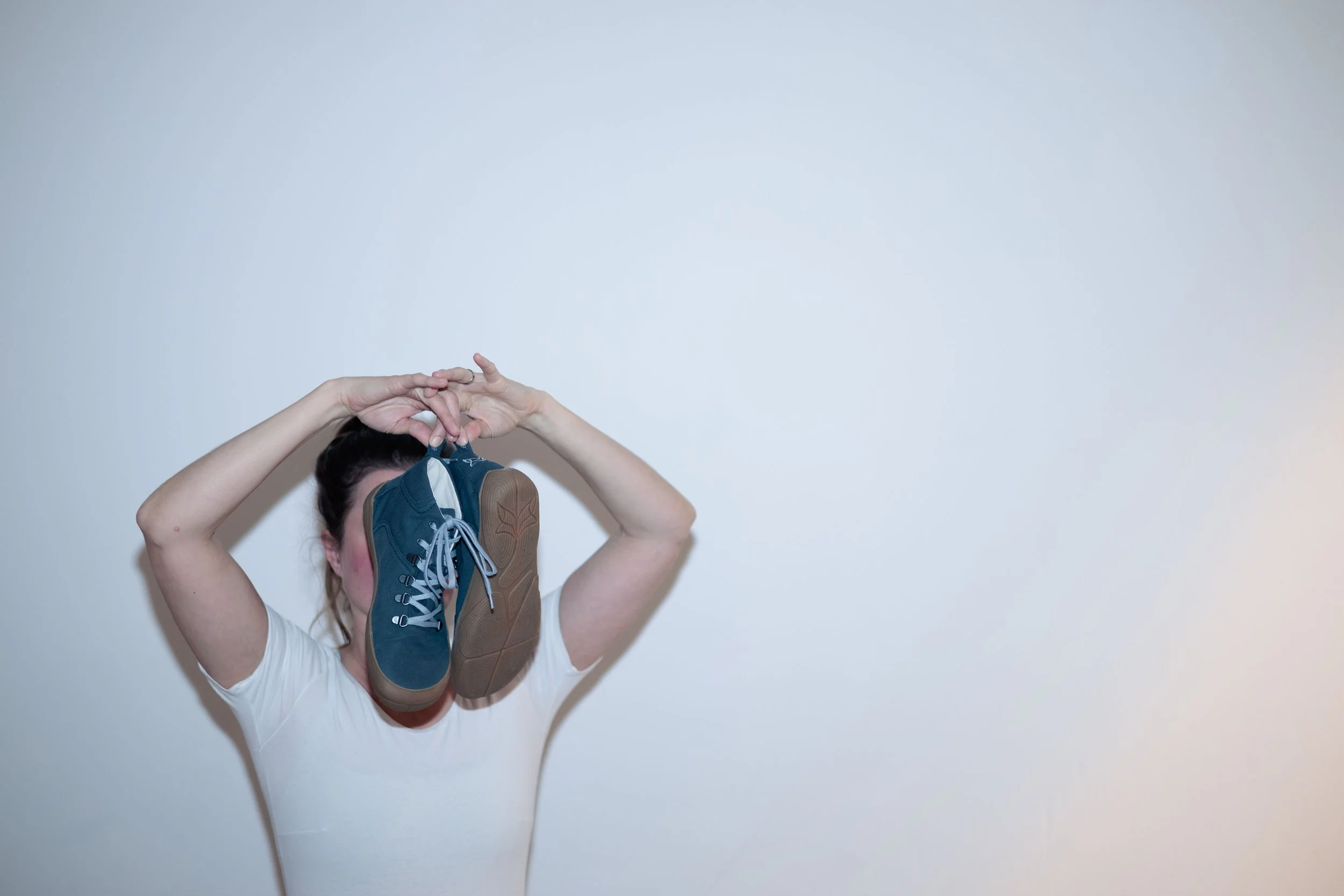 Fashion photography München. Frau steht vor einer weißen Wand und hält sich ein paar Schuhe vor das Gesicht.