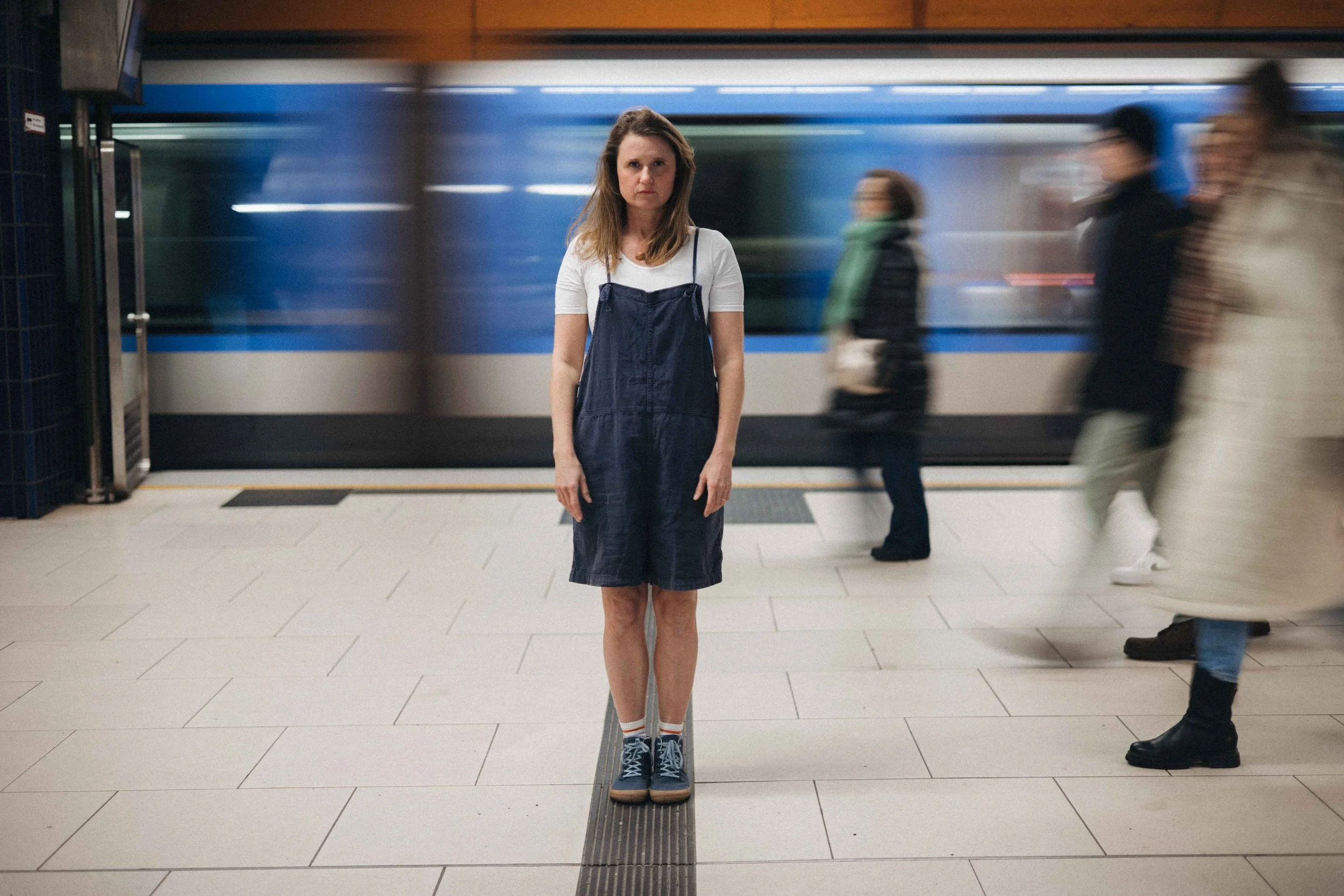 Editorial Fotoshooting in Münchner U-Bahn Station. Frau steht still und um sie herum bewegen sich Menschen. Im Hintergrund fährt eine Bahn vorbei.