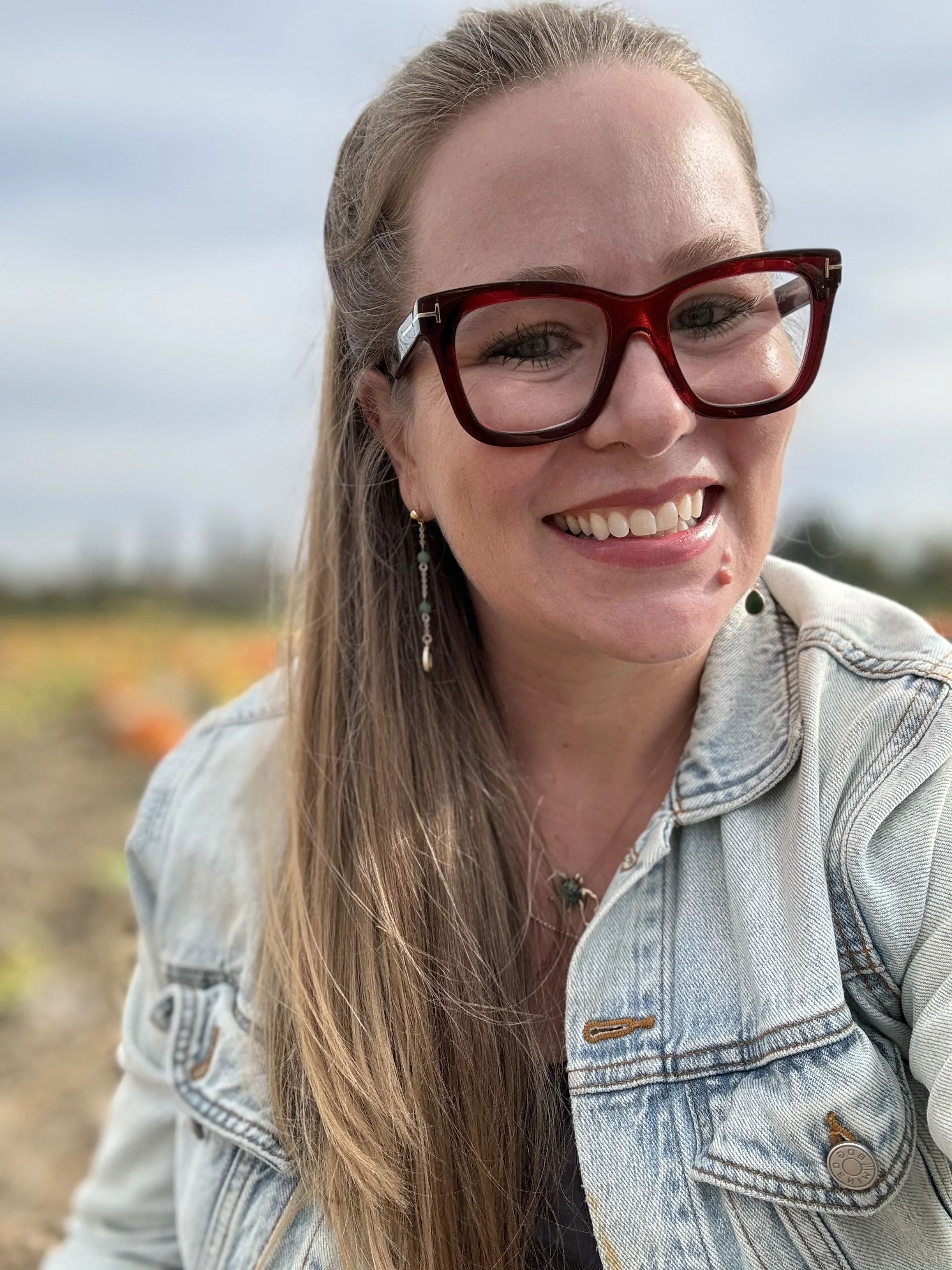 Leslie Bradshaw wearing Tom Ford red glasses and Old Navy green earrings, with long light brown hair, outdoors in Oregon a field with pumpkins and a cloudy sky in the background.